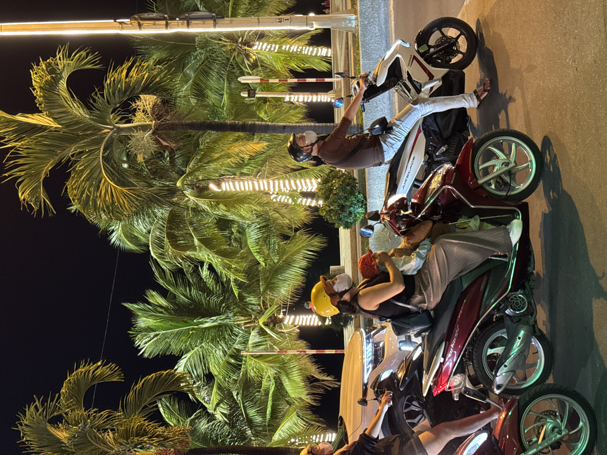 The image depicts a nighttime scene in Nha Trang, Vietnam, likely on a street lined with palm trees decorated with string lights. Several people are riding on motorcycles. In the foreground, a woman wearing a yellow helmet and a mask is holding a baby, who also wears a mask and a red hat. They are on a red motorcycle. To the right, a man in a brown shirt and a helmet, also wearing a mask, rides a white and black motorcycle. Further to the left, a person wearing a yellow helmet and a mask is on a motorcycle, partially obscured by a white car. The overall atmosphere suggests a typical urban evening in a tropical setting.
