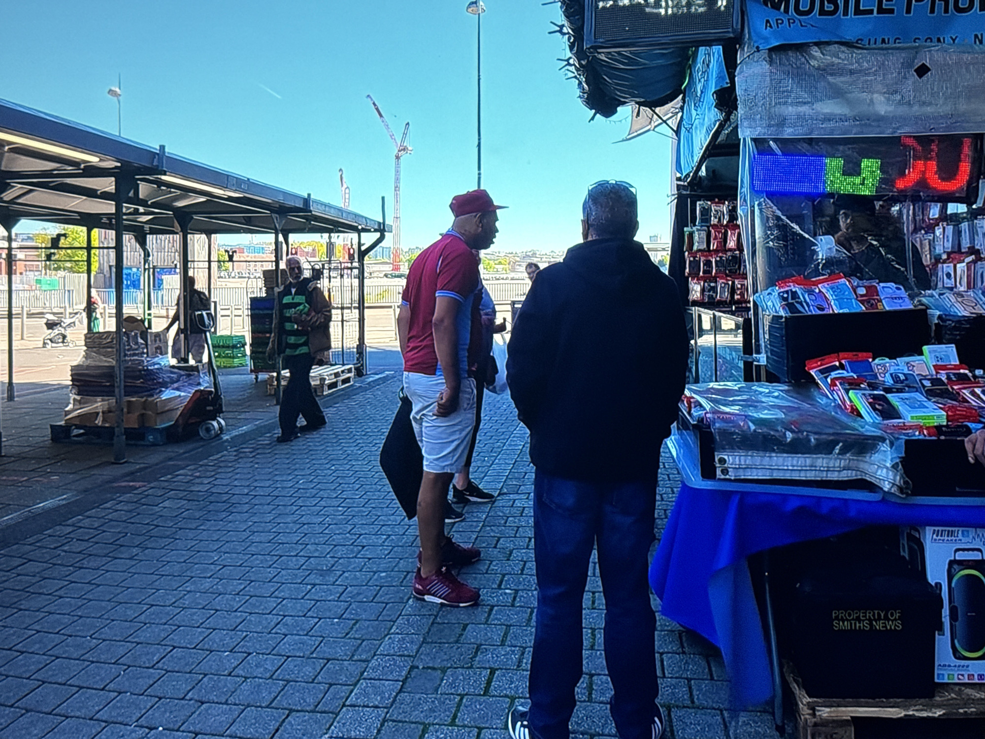 The image depicts an outdoor market scene. The foreground is dominated by a cobbled street. In the midground, several individuals are visible. Two men, one wearing a maroon and blue football jersey and white shorts, and another in a dark jacket and blue jeans, are facing away from the camera, appearing to converse. To the left, another person in a dark jacket stands near a covered stall. Further left, a person is pushing a pallet jack loaded with boxes under a covered structure.
In the background, the market stalls continue. One stall prominently displays various mobile phone accessories. A blue banner above the stall reads "MOBILE PH". A neon sign displays "JU" in red and blue lights. Beyond the stalls, a fence and some buildings are visible. In the distance, construction cranes are present against a clear blue sky. The overall impression is a bustling market on a sunny day, likely in an urban or semi-urban environment. The specific location of Cheltenham, United Kingdom, is not directly evident from the visual information in the image.