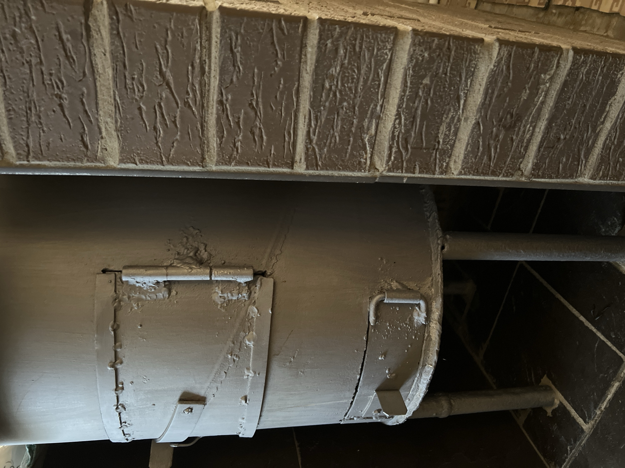 The image shows a close-up, medium-angle shot of an old, gray metal stove positioned next to a brick wall. The stove is cylindrical with visible seams, rivets, and hinges. It appears to be supported by multiple legs. There are two doors on the stove, one larger and one smaller near the bottom, both with handles. The paint on the stove is chipped and worn in several places, showing signs of age and use. The brick wall to the right has a textured surface with horizontal lines, suggesting it is made of rough-hewn bricks. The floor beneath the stove is tiled with dark, square tiles, separated by light-colored grout. The lighting suggests an indoor environment, possibly a room with minimal light. There are no people or animals visible in the image. The scene is static and offers no indication of ongoing activity. The context of Khryashchevka, Russia, is not visually present in the image itself.