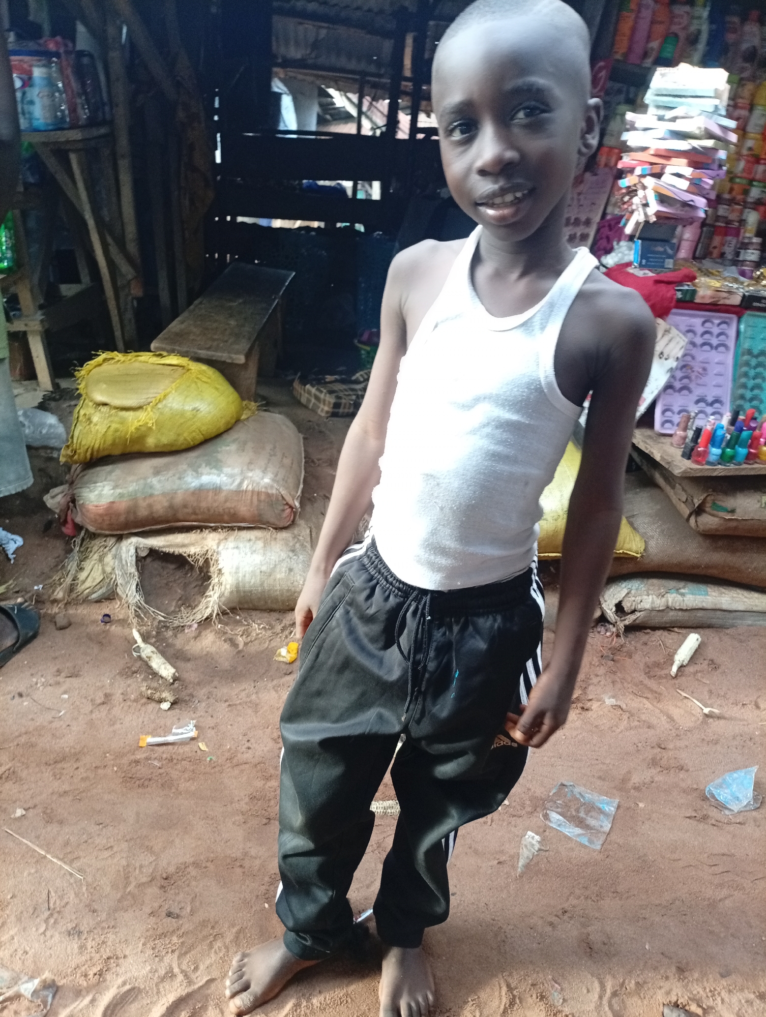 A young boy, with a close-cropped haircut and a bright smile, stands barefoot on a sandy, dirt ground in Nkpor, Nigeria. He wears a white tank top and black athletic pants with white stripes down the sides. Behind him, a bustling market scene unfolds. Stacks of colorful products, likely cosmetics and toiletries, are displayed on wooden shelves. Several large sacks, one bright yellow, are piled on the ground, suggesting goods for sale or storage. The background shows makeshift structures with corrugated metal roofing, indicating a typical market environment. The lighting suggests it could be daytime, with the sun likely overhead given the shadows. The boy's expression is one of joy and confidence, as he looks directly at the camera. The overall impression is a snapshot of everyday life in a Nigerian market.