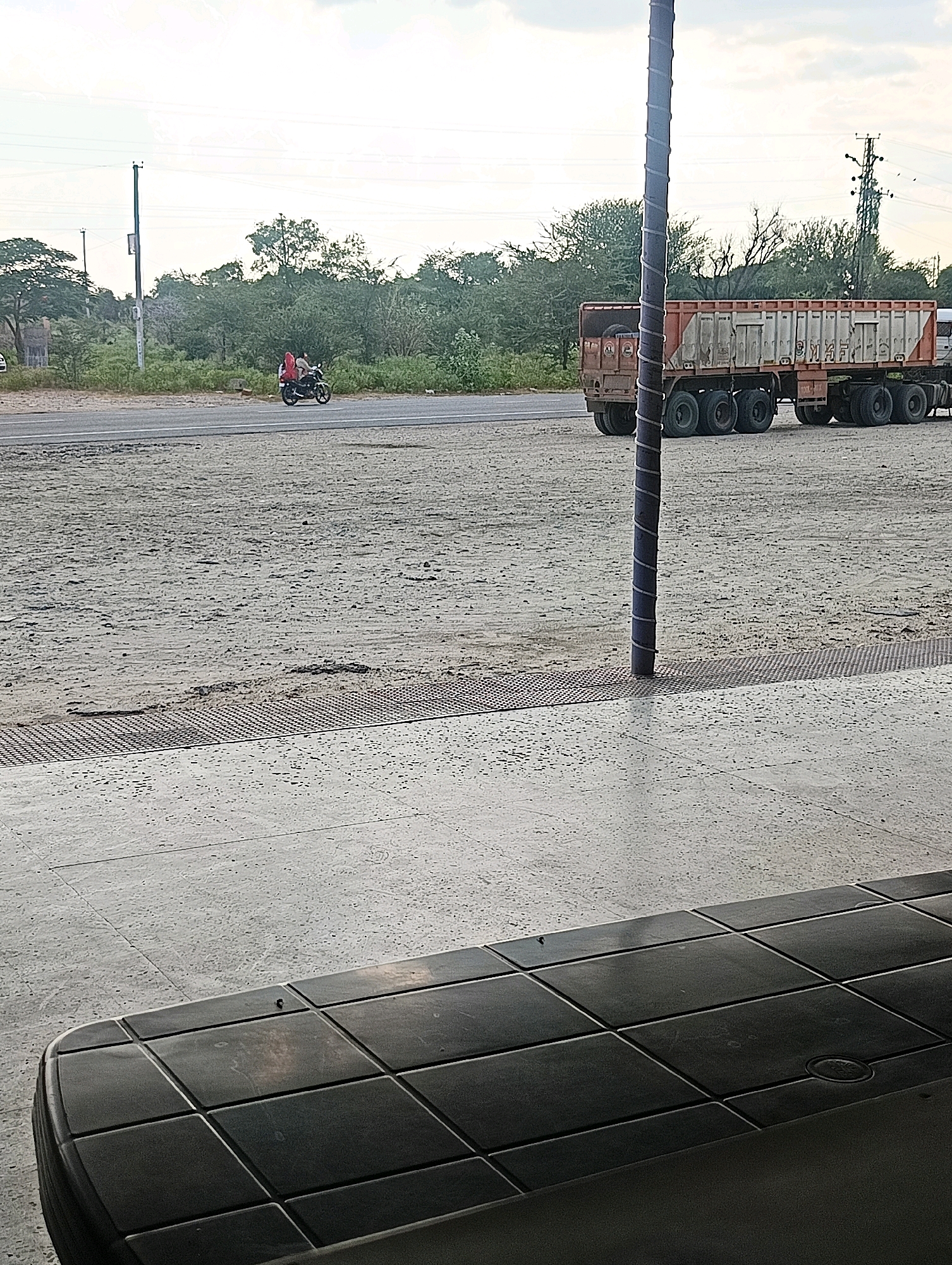 In Chhapri Bari, India, a motorbike with two occupants, a driver and a passenger, travels on a paved road. They are both wearing red garments. Beside the road, a large truck with an orange and white trailer is parked on a gravel lot. In the foreground, a section of a dark, segmented surface, possibly a table or bench, is visible, with a textured concrete ground in front of it. The background shows a line of trees and utility poles against a cloudy sky.