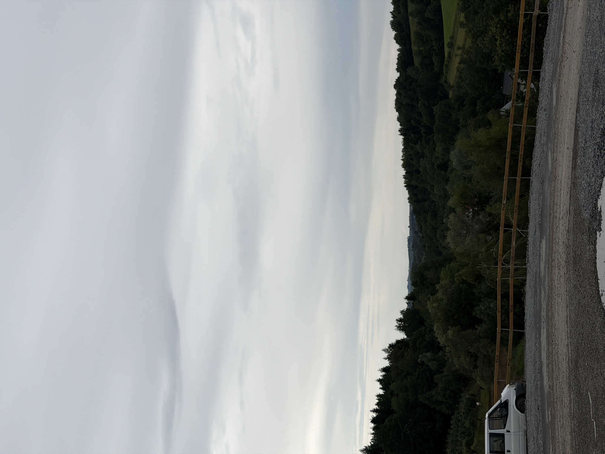 This media file depicts an outdoor scene at dusk, likely in Stróża, Poland. The image is oriented vertically, showing a vast expanse of pale, overcast sky on the left, transitioning to a dense, dark forest on the right. A wooden fence with horizontal slats separates the forest from a gravel path. A white van is parked on the gravel path. The overall mood is peaceful and serene, with the soft, diffused light of dusk contributing to the calm atmosphere. The weather appears to be overcast, with no visible sun. The lush green of the forest suggests a temperate climate. The presence of the van and fence indicate a human presence and potentially a rural or semi-rural setting. There is no visible text in the image.