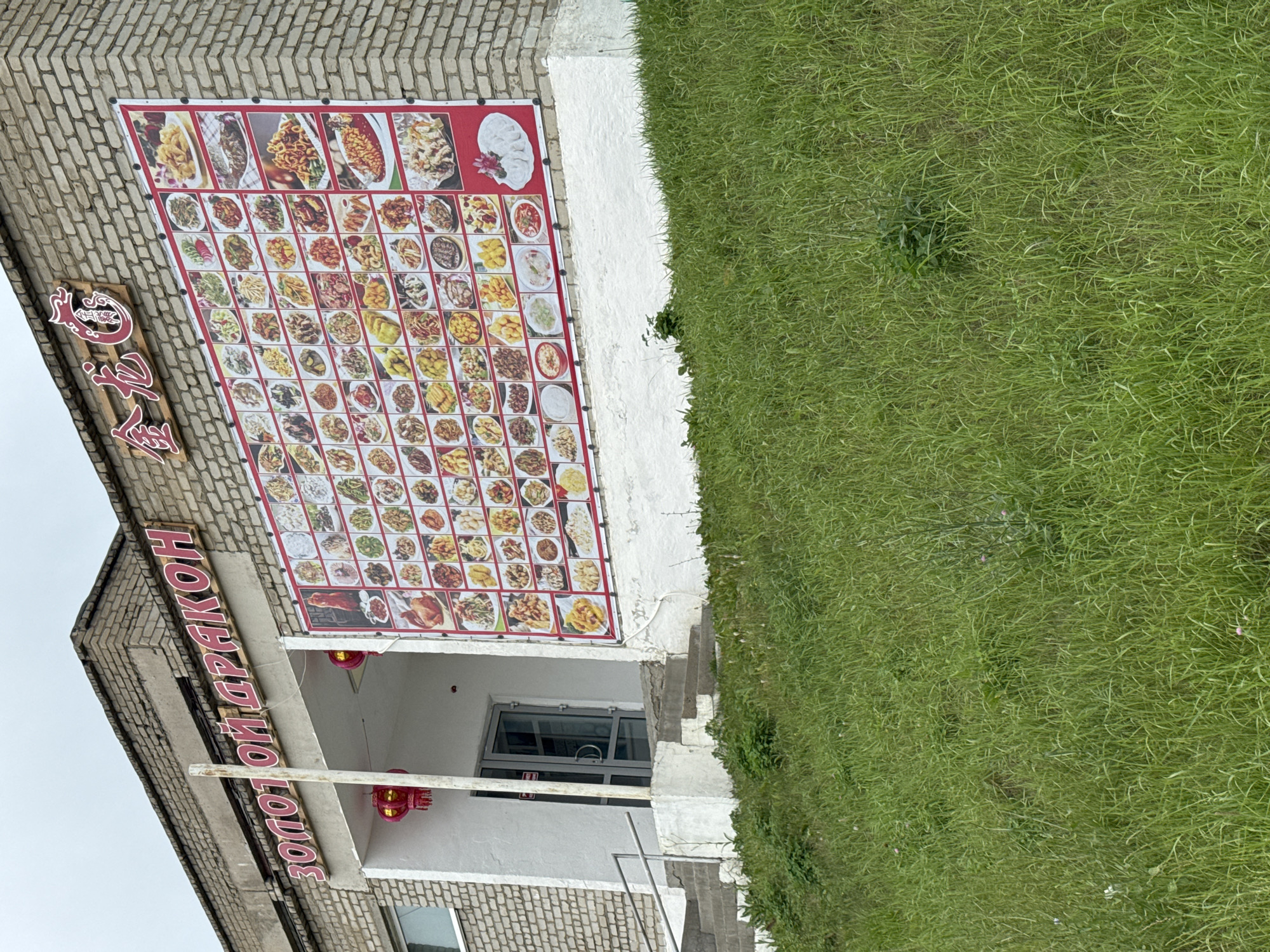 The image shows the exterior of a Chinese restaurant named "ЗОЛОТОЙ ДРАКОН" (Golden Dragon) in Chuguyevka, Russia. The building is made of brick. A large banner displaying various Chinese dishes is mounted on the exterior wall. There is a glass door entrance with a set of stairs leading up to it. Red lanterns are hanging near the entrance. The restaurant is situated on a grassy slope.