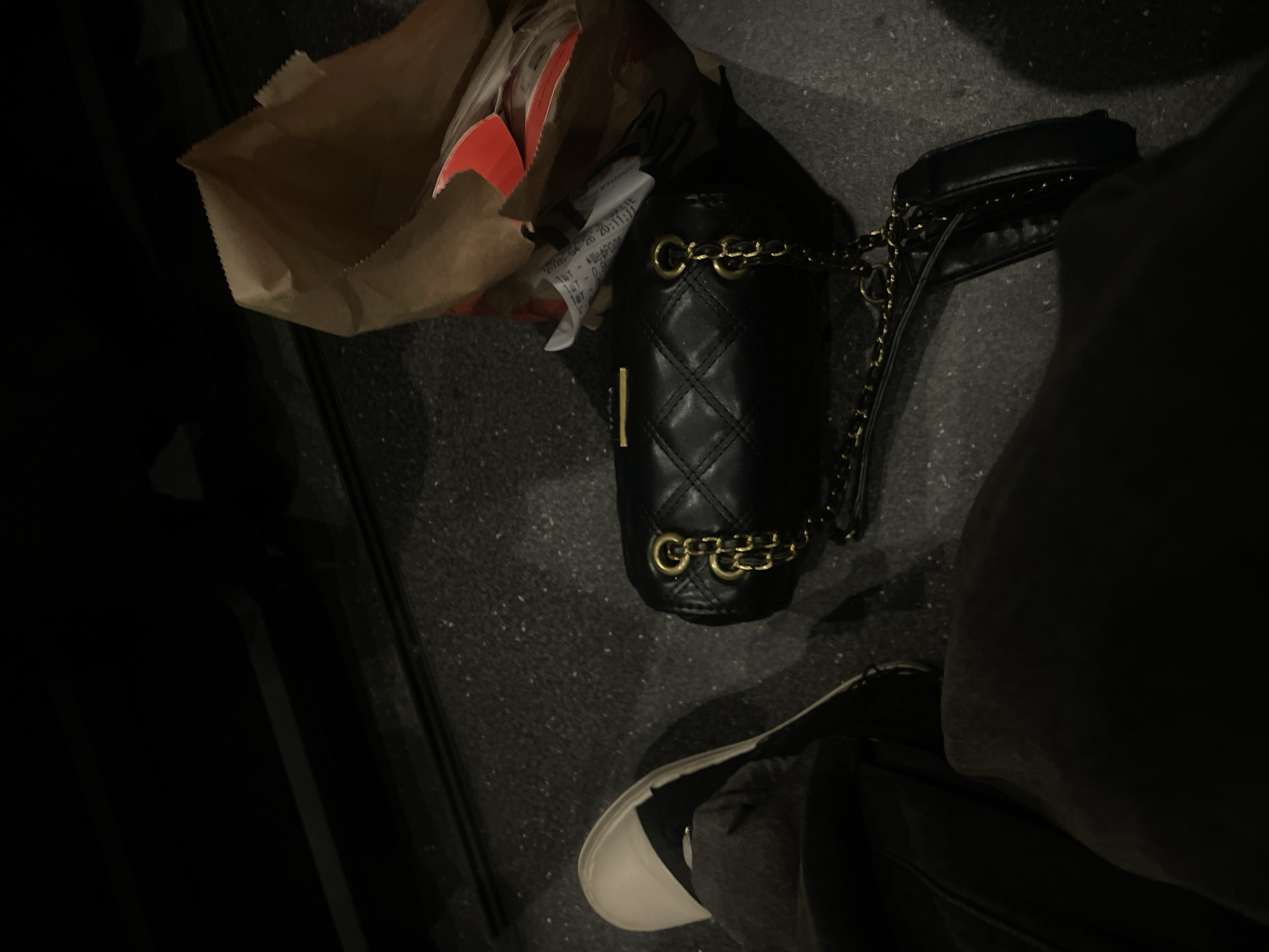 The image shows a black quilted handbag with a gold chain strap lying on a dark grey surface, possibly a floor or street. Next to the bag is a brown paper bag with a fast food receipt sticking out. The bottom of a person's leg and shoe are visible near the bottom right of the image. The shoe has a white toe and black trim. The image is somewhat dark, making it difficult to discern finer details. Location cannot be determined.