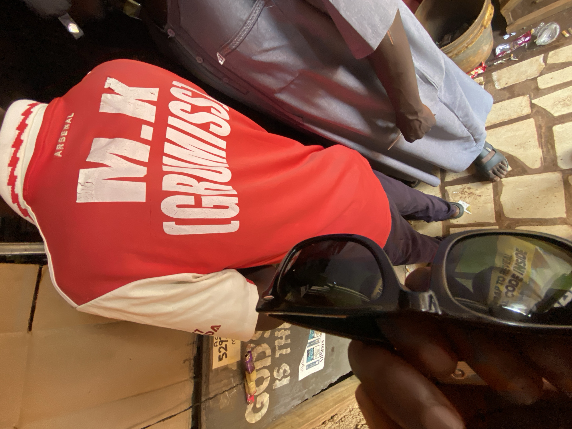 In Gura Top, Nigeria, an individual wearing a red and white Arsenal football jersey with "M.K. GRUMISS" and "ARSENAL" printed on the back is captured from the upper torso down. The jersey has a white collar with a red and white zig-zag pattern. The person is also wearing dark trousers and dark sandals. In the foreground, a hand holds a pair of black sunglasses, reflecting a yellow label that reads "SCRAP TO REVEAL CODE INSIDE". Behind the person in the jersey, another individual is partially visible, wearing a light grey garment and dark sandals. The environment appears to be outdoors, with textured walls and floor tiles. A sign with "GOD'S IS TH" and numbers is visible on the wall. Various objects, including candy wrappers and what appears to be a large metal drum, are scattered around the ground.