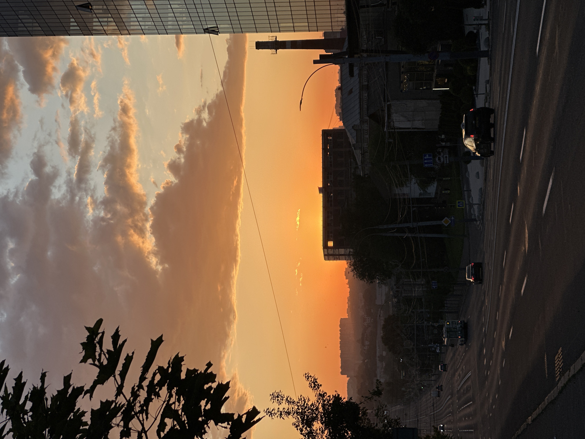 The image captures a sunset over a city street, likely in Chișinău, Moldova, given the context. In the foreground, a dark road stretches uphill with lane markings. Several cars, including a dark sedan and a trolleybus, are visible on the road. On the right side of the street, a modern glass-faced building stands tall. Further back, the silhouette of an unfinished building with visible beams is present, alongside a tall industrial chimney. The sky is a vibrant orange and yellow, with scattered clouds, indicating the sun is low on the horizon. Silhouetted trees are present on the left, with dark leaves forming a frame at the top left. The overall scene depicts an urban environment during twilight.