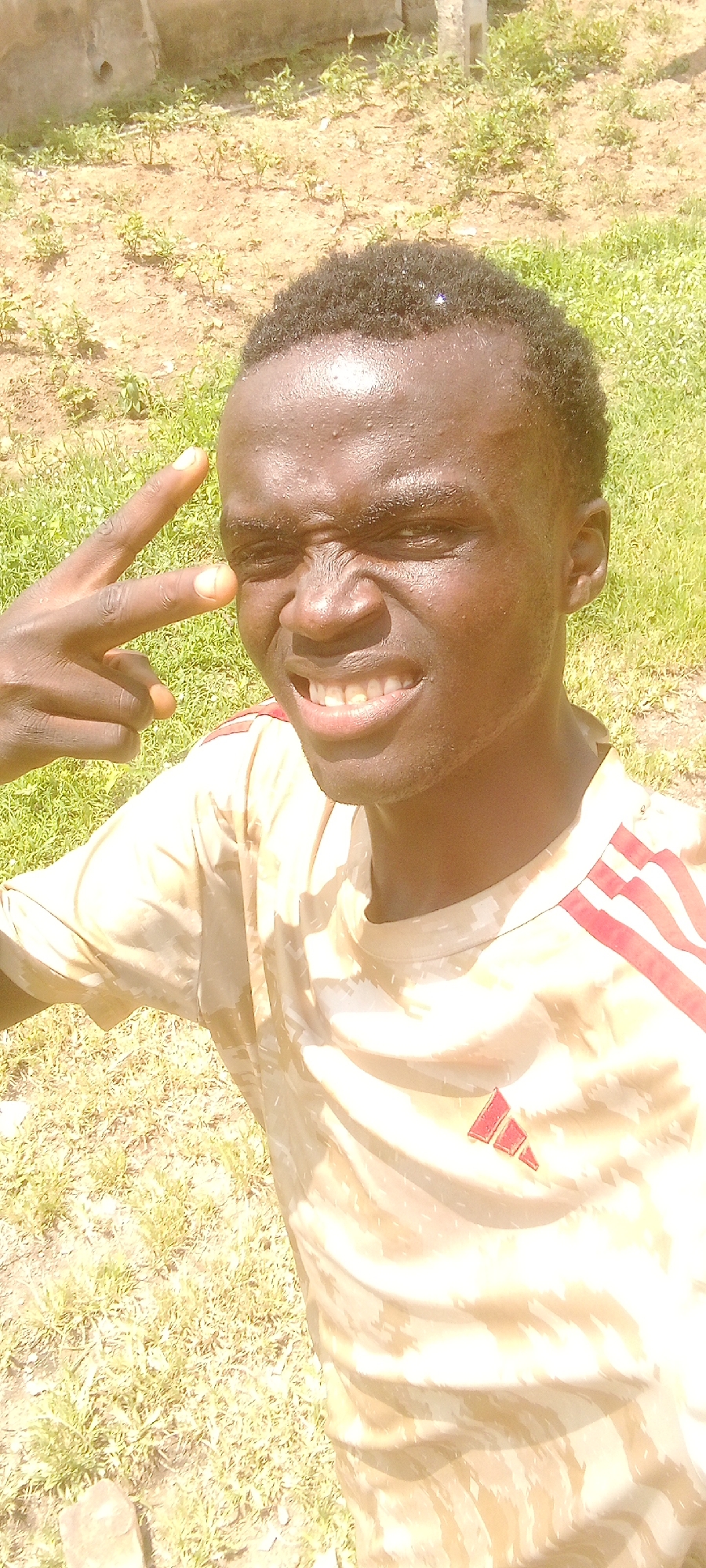 A young Black man with dark skin and short, dark hair is shown in a close-up, outdoor selfie. He is wearing a light-colored t-shirt with a camouflage-like pattern and red stripes on the shoulders, along with an Adidas logo. His face is contorted in an exaggerated expression with his eyes squinted and his mouth open in a grimace, revealing his teeth. He is holding up two fingers to his forehead in a gesture that resembles a gun. The background is blurred and appears to be a mix of dry dirt and green grass, with some sparse vegetation. The lighting suggests it is daytime and sunny, with some overexposure.