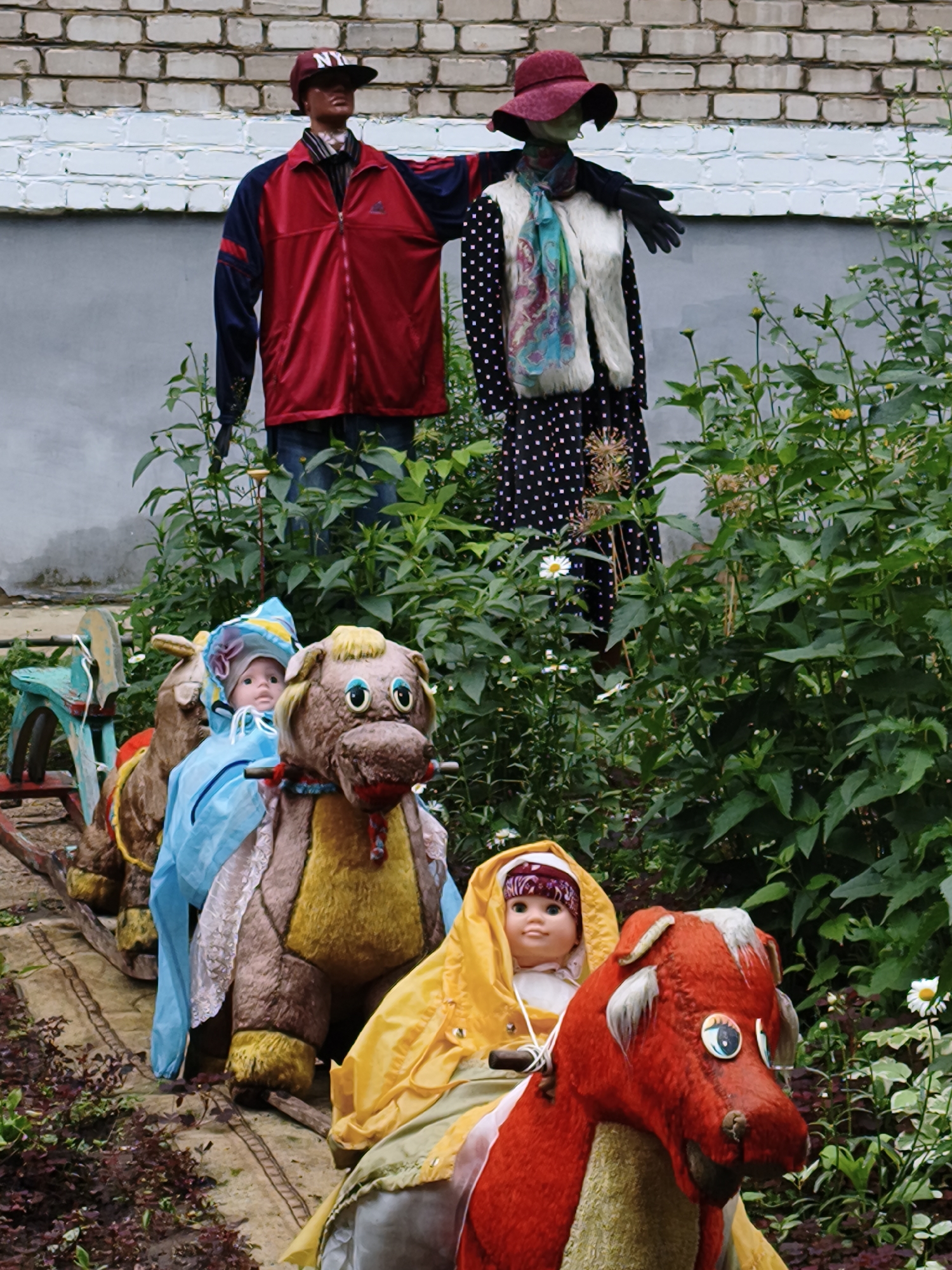 In Chaikovsky, Russia, two mannequins stand dressed as if for a cooler climate. The one on the left wears a red and navy jacket, blue jeans, and a red baseball cap. The mannequin on the right is adorned in a black and white polka dot dress, a light-colored vest, a colorful scarf, and a wide-brimmed red hat. In front of them, on a miniature train track, several stuffed animals are positioned, resembling a carousel ride. A doll sits atop a red and tan creature, draped in a yellow raincoat. Another doll in a blue hooded garment is seated on a brown, horse-like stuffed animal. A third, mostly obscured, is on a brown creature with a weathered blue rocking horse in the background. The scene is set against a backdrop of a brick wall and lush green foliage.
