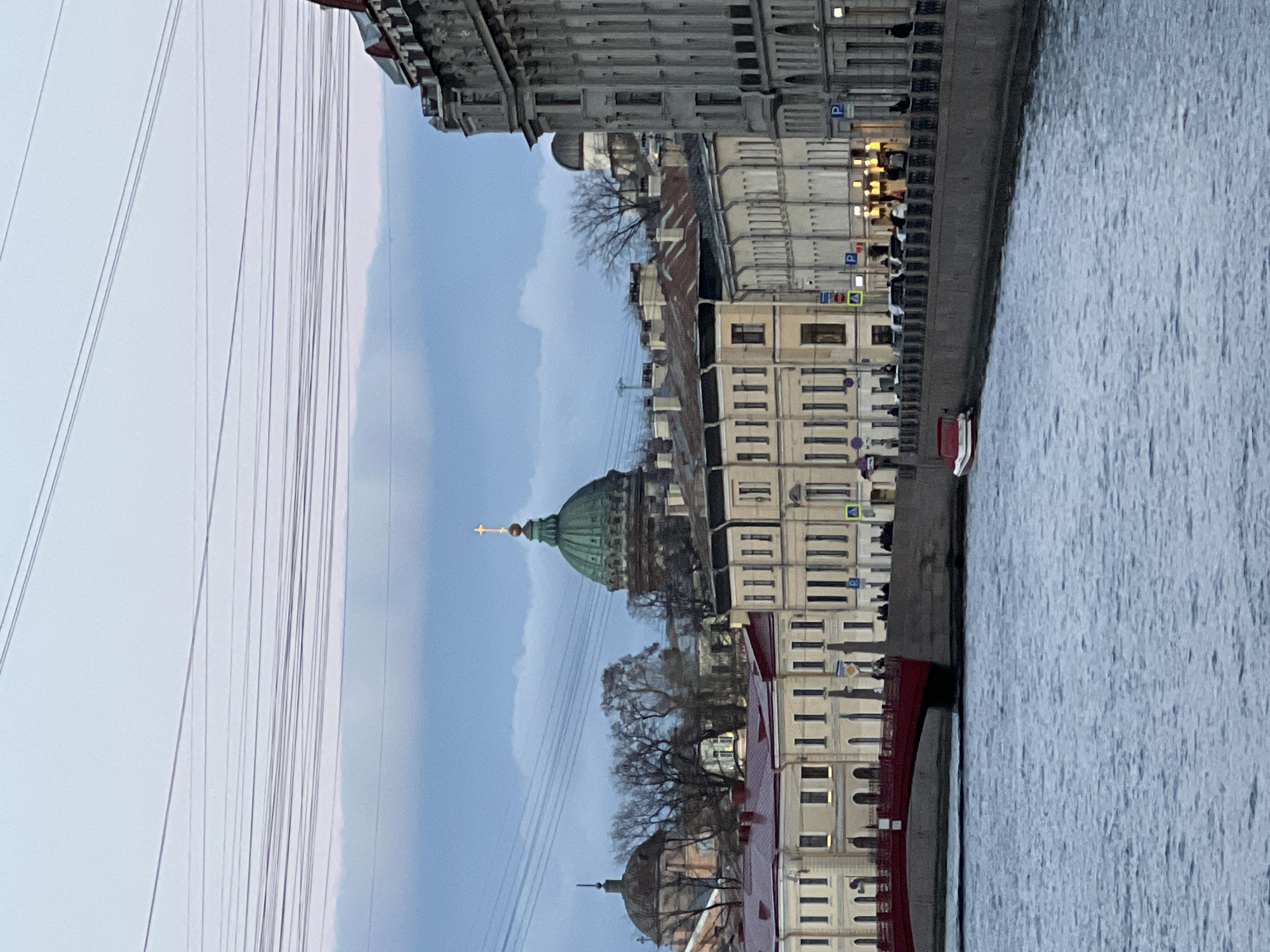 The image shows a waterfront scene in Saint Petersburg, Russia. The iconic green dome of St. Isaac's Cathedral is visible, situated amongst other buildings with pale facades. A body of water, likely the Neva River or a canal, is on the right of the frame. Some people are along the waterfront. Overhead power lines are present. The sky is overcast.