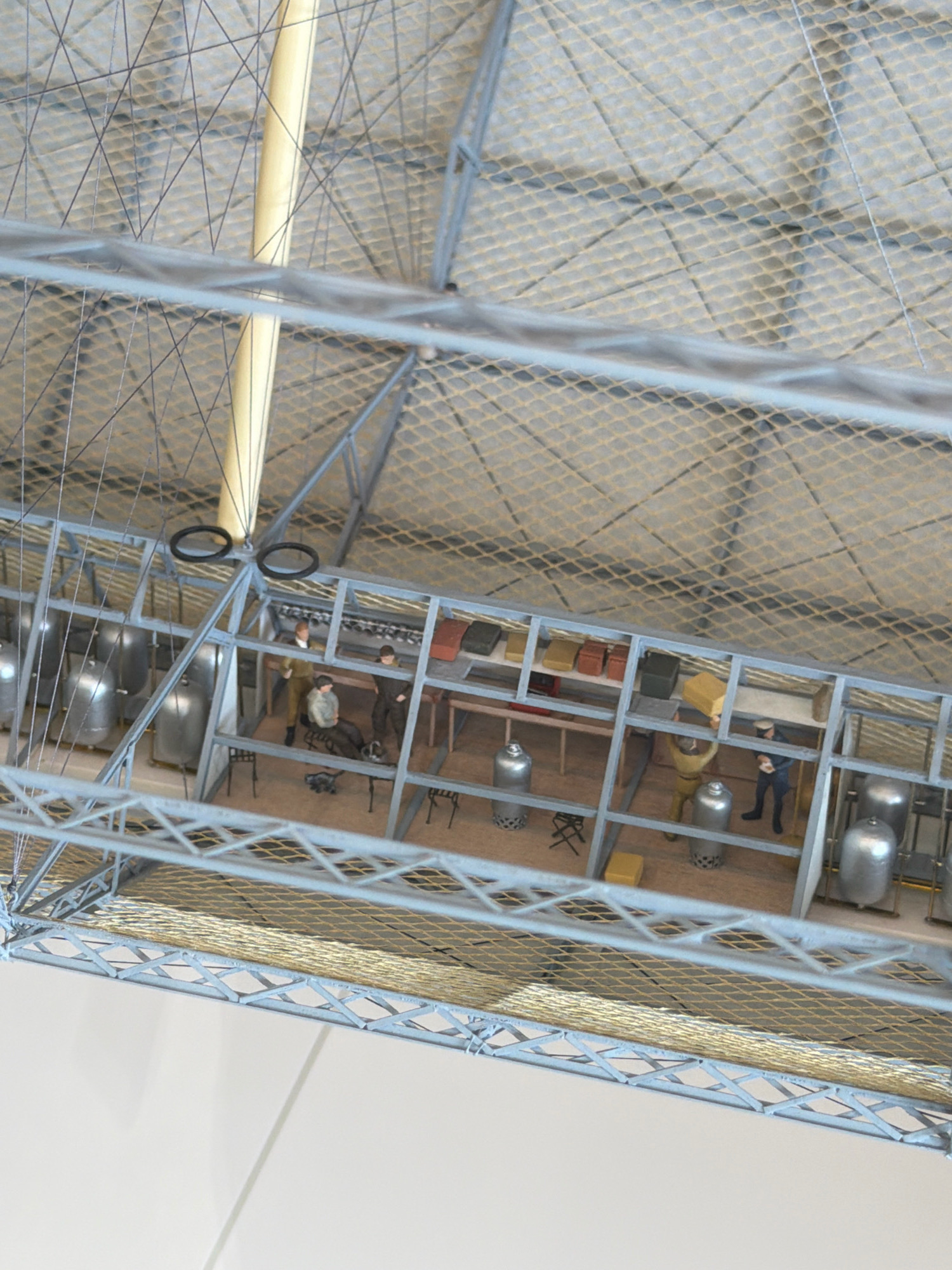 This is a detailed model of the interior of the Zeppelin hangar at Friedrichshafen, Germany, showcasing a scene within a workshop or laboratory. The model depicts a structured environment with a metal framework supporting a meshed ceiling. In the foreground, a section of the hangar's supporting structure is visible, constructed from light-colored metal beams and cross-bracing.
Further inside the model, a long, narrow workshop area is represented. This space is divided into several compartments by vertical metal supports. The flooring appears to be made of wood.
Several metallic cylindrical tanks, some with textured bases, are arranged within the workshop. These tanks are likely intended to represent storage or processing vessels.
There are several miniature figures, representing people, engaged in various activities. In one section, two figures are near what appears to be a workbench or shelving unit with colorful boxes. Another figure is seated near a larger cylindrical object. Further down the workshop, two figures are interacting near a table, one holding what looks like a document or plan. A small, dark, four-legged object, possibly a miniature dog, is also present on the floor near the seated figure.
The lighting within the model suggests an indoor environment, possibly with overhead illumination. The overall scene conveys a sense of industrial activity or scientific work taking place within the historical context of the Zeppelin era in Friedrichshafen.