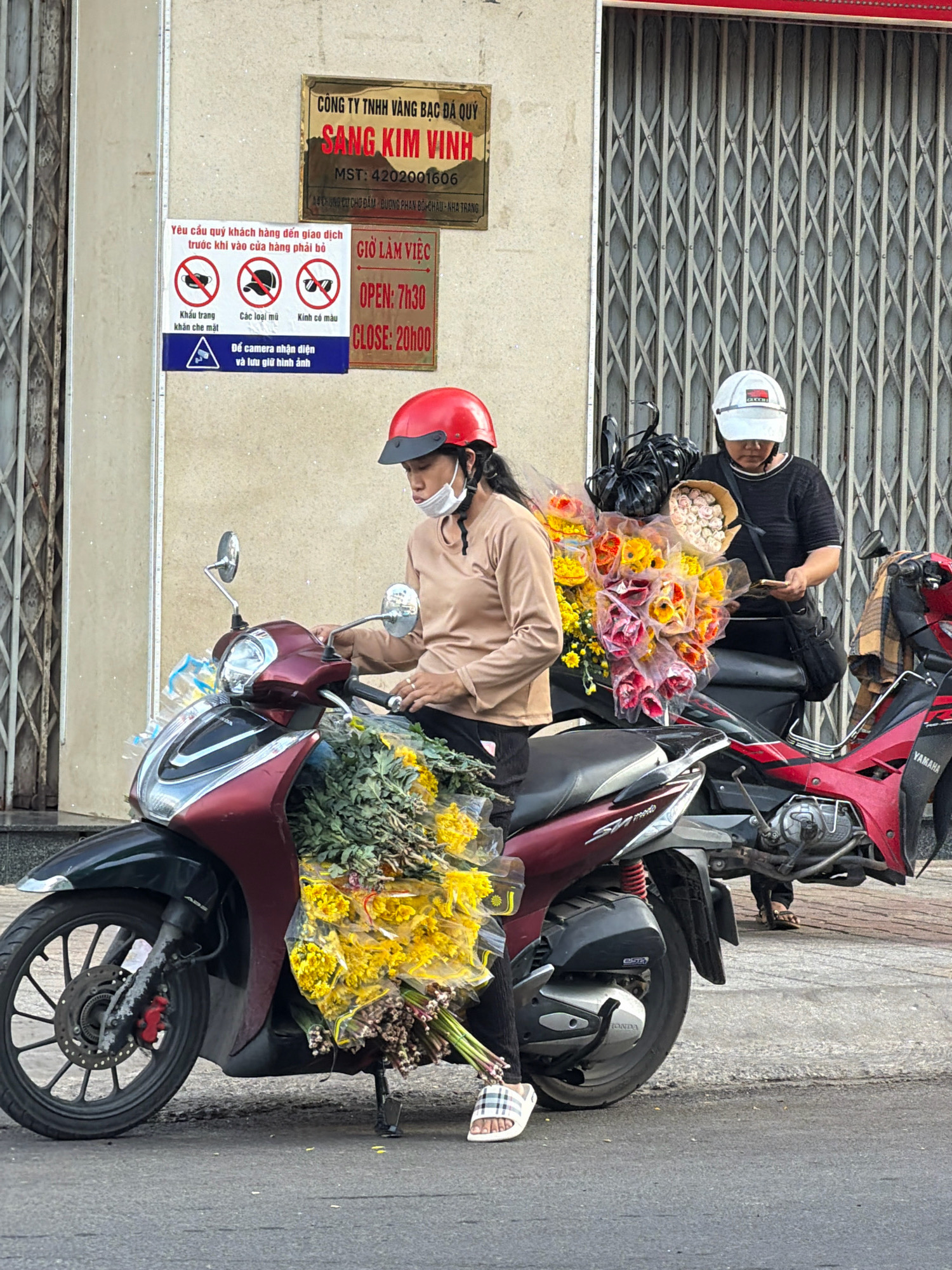 The image depicts a street scene in Nha Trang, Vietnam, with two individuals on motorcycles laden with flowers. In the foreground, a woman wearing a red helmet and a face mask is seated on a maroon scooter. She is dressed in a light brown long-sleeved shirt and black pants, with a white and checkered sandal on her right foot. A large bunch of yellow and green flowers is prominently displayed on the scooter in front of her. Behind her, a second person wearing a white baseball cap and a black shirt is on a red motorcycle. This individual appears to be looking at a mobile phone. Both motorcycles are parked on a paved street with a metallic accordion-style gate in the background. Signs with Vietnamese text are affixed to the wall beside the gate, including one indicating business hours and another with cautionary symbols. The overall setting suggests a commercial area where flowers are being sold or transported.