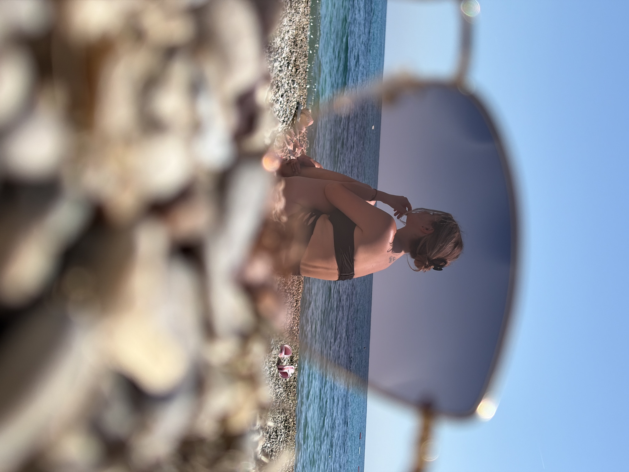 A person wearing a dark-colored bikini is sitting on a pebble beach next to the sea, as viewed through sunglasses. Two pink sandals sit on the pebbles near the water's edge. The sky is blue and clear. The foreground is filled with blurry pebbles. Location: The location is Prijevor, Montenegro.