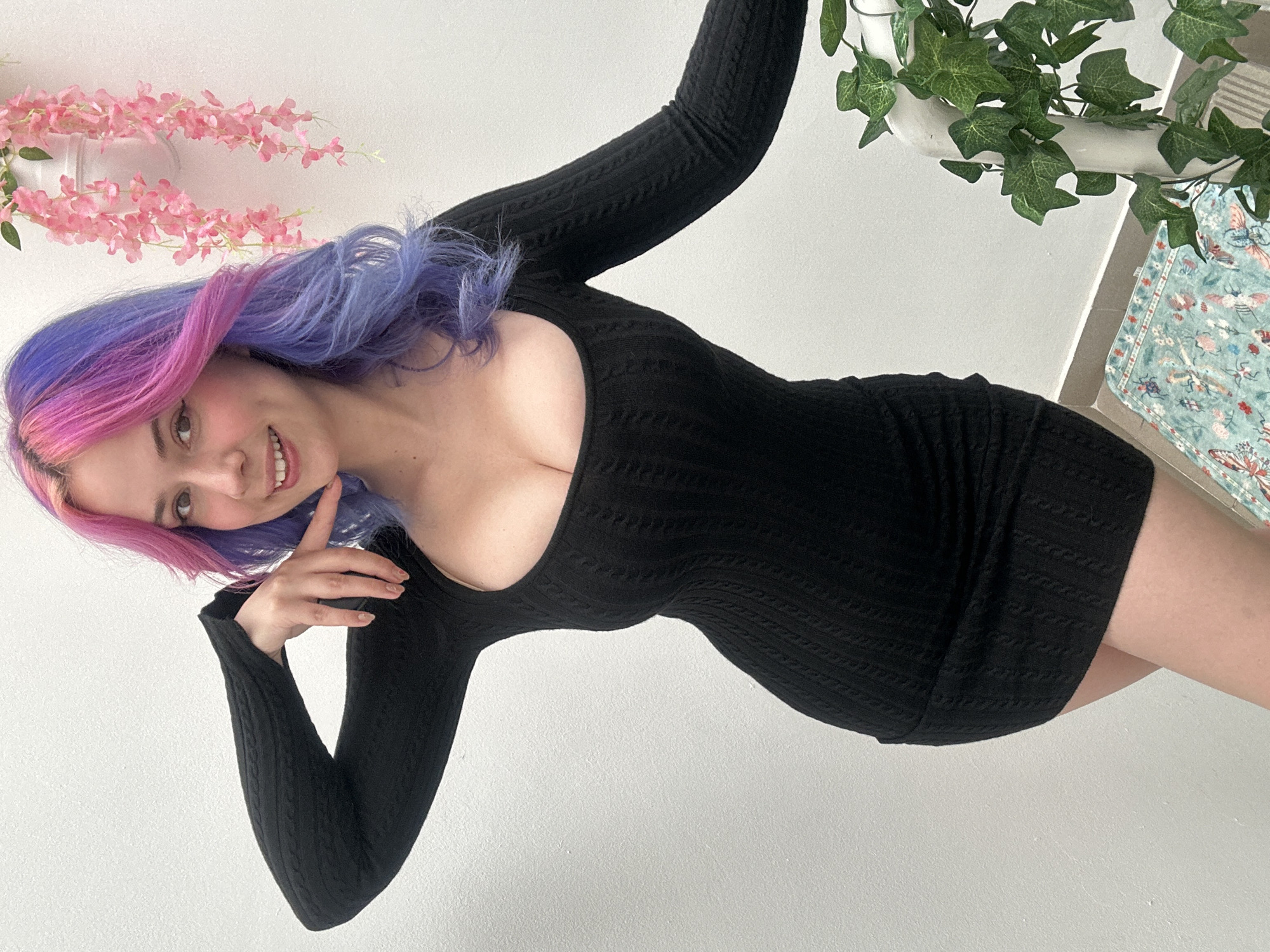 The image captures a young woman with distinctive, long hair dyed with an ombre effect, transitioning from bright pink at the roots to light purple at the ends. She is smiling widely at the camera, her head tilted, and one hand gently resting near her chin. She wears a form-fitting, black long-sleeved dress with a scoop neckline and a subtle, textured knit pattern.
The subject is positioned in a way that suggests she is leaning or reclining, with the background appearing tilted. The backdrop is predominantly a plain white wall. To the left, a decorative garland of artificial pink flowers hangs. To the right, artificial green ivy is draped, and a small section of light blue fabric with a patterned design (possibly butterflies or insects) is visible. The lighting is bright and even, consistent with an indoor setting. The woman's expression conveys happiness and friendliness. There is no visible text. The provided location context for the image is the City of Zagreb, Croatia.