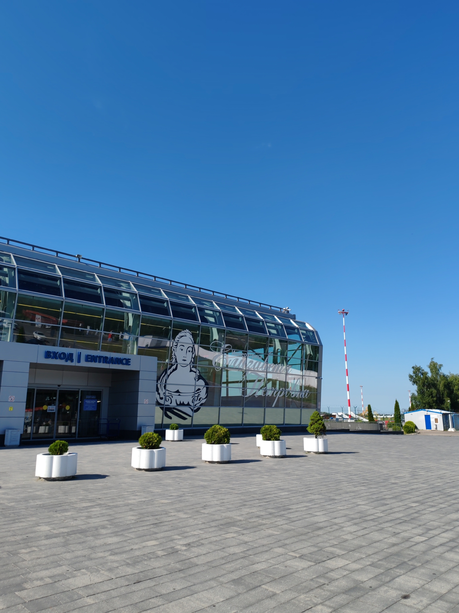 The media file depicts the exterior of an airport terminal building on a clear, sunny day. The building features a modern design with a large glass facade that reflects the bright blue sky and surrounding environment. A prominent black and white graphic of a woman's bust is emblazoned on the glass. The entrance to the building is clearly marked with the word "ВХОД" (Entrance) in blue lettering, followed by "ENTRANCE" in English.
In front of the building, a paved plaza is lined with white planters, each containing a neatly trimmed green bush. Beyond the building, a red and white striped pole, likely an airport indicator, is visible against the sky. Further in the distance, trees and a small blue and white building can be seen. The overall scene suggests a well-maintained and functional airport setting under good weather conditions. The time of day appears to be daytime, given the bright sunlight and clear sky. No people are actively engaged in any activity within the frame, giving the scene a quiet, perhaps early morning or late afternoon, feel. The location is identified as Khrabrovo, Russia.