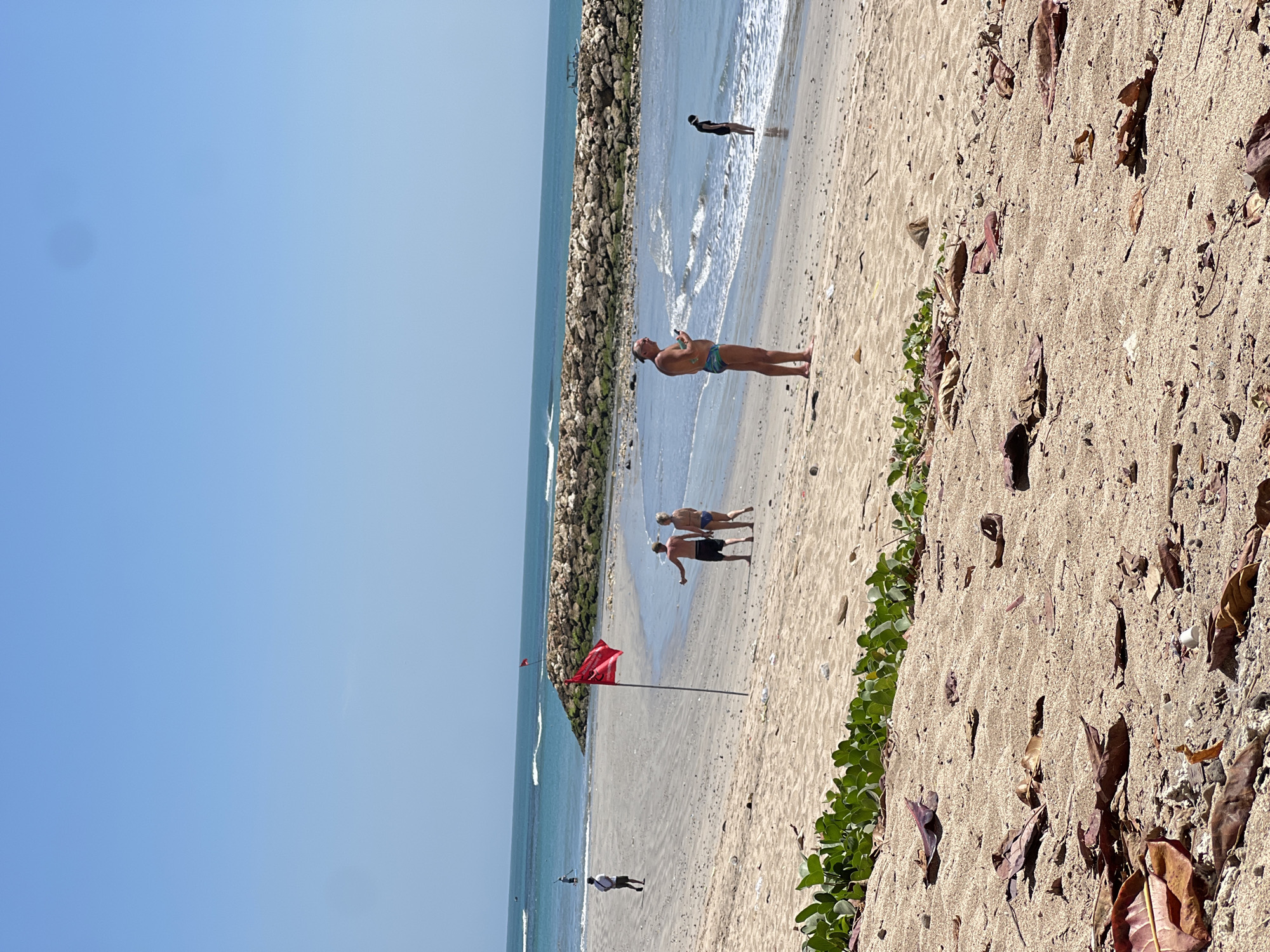 The image depicts a beach scene in Kuta, Indonesia, under a clear blue sky. The foreground is dominated by sandy beach with scattered fallen leaves and some greenery. In the midground, a red flag flies on a pole, positioned on the sandy shore. A couple, a man and a woman, are walking away from the camera, towards the water, with the man pointing to something in the distance. To the left of them, another man is walking on the beach. Further out, several people are visible near the water, and a stone breakwater runs parallel to the shore. The ocean has gentle waves. In the distance, a boat is visible on the horizon.