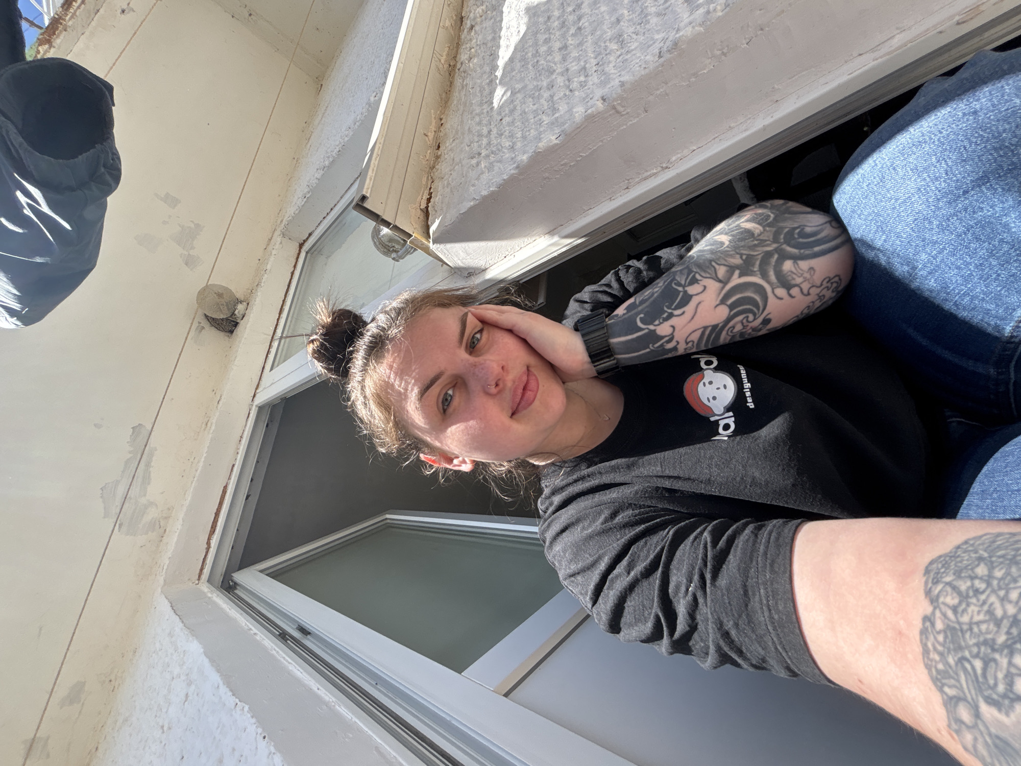 A person is leaning out of a window. The person has a bun hairstyle, tattoos on their arms, and is wearing a black long-sleeved shirt and blue jeans. The window appears to be in a building with peeling white paint. There is a dark object hanging outside the window to the left. The exact location in Chornomorskoe, Ukraine, cannot be determined based on the visual information.