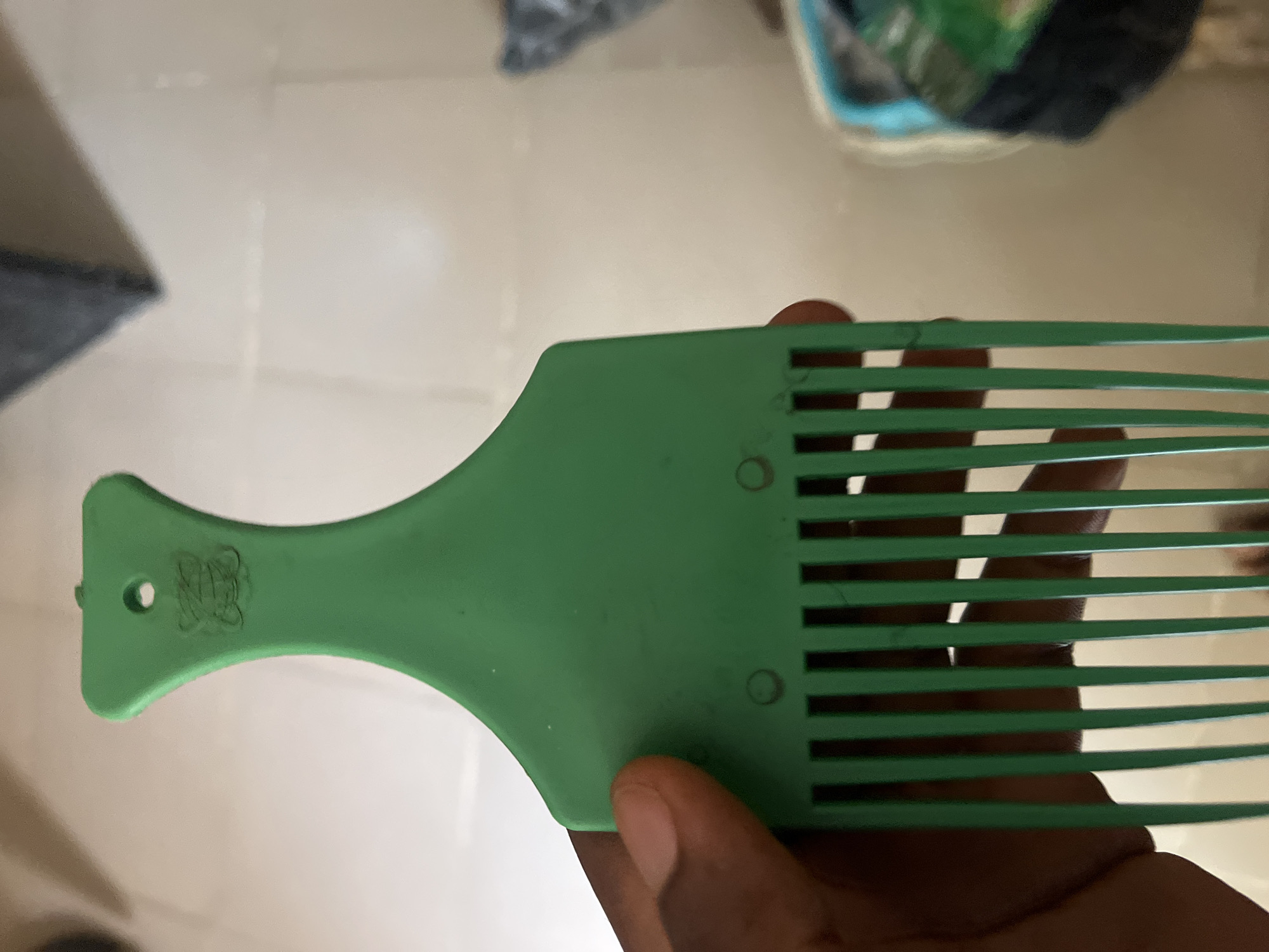 A green plastic afro pick is held by a person's hand. The pick has a wide handle with a small hole at the top and a decorative logo embossed on the handle, consisting of what appears to be interwoven letters. The lower part of the pick features widely spaced teeth. The background is blurred but suggests an indoor setting with light-colored walls and a floor, possibly in Jalingo, Nigeria.