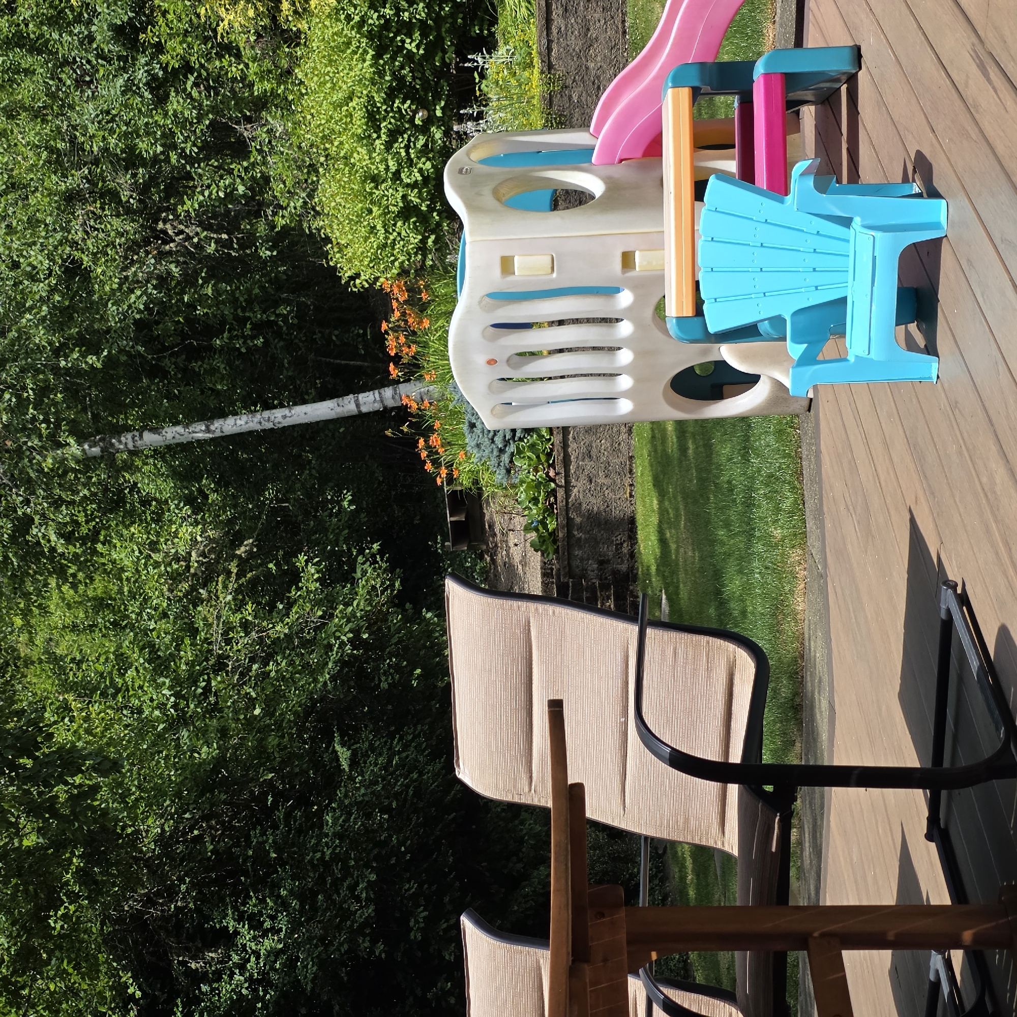 A bright, sunlit backyard scene unfolds on a wooden deck in Portland, Oregon. In the foreground, two large, neutral-toned outdoor chairs with striped cushions are positioned next to a wooden dining table. To the right of the chairs, a child's play structure featuring a white and blue climbing tower with a pink slide is visible. A small, bright blue plastic Adirondack-style chair sits in front of the play structure. The background is filled with lush green foliage, including trees and bushes, with a cluster of vibrant orange flowers adding a pop of color. The overall impression is one of a well-maintained outdoor living space ready for recreational activities.