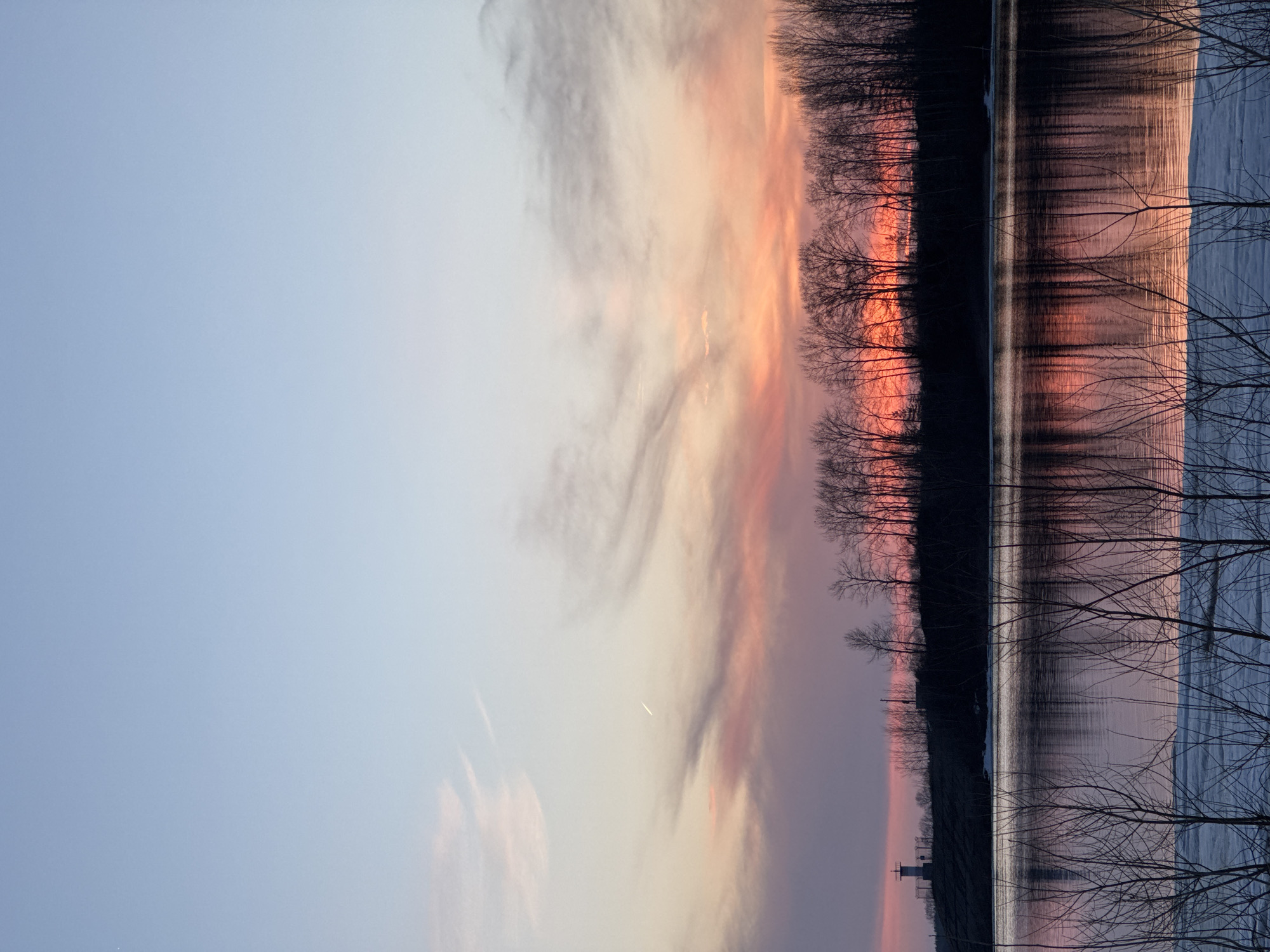 The image depicts a sunset scene over a body of water, possibly in Chaikovsky, Russia, based on the location request. The sky is filled with streaks of orange, pink, and purple hues blending with the pale blue of the early evening sky. A line of dark trees silhouettes against the colorful sunset. Reflections of the sky and trees are visible in the water. Bare branches extend from the right side of the frame. There are no people visible in the image. A small structure can be seen at the edge of the water, potentially a lighthouse.