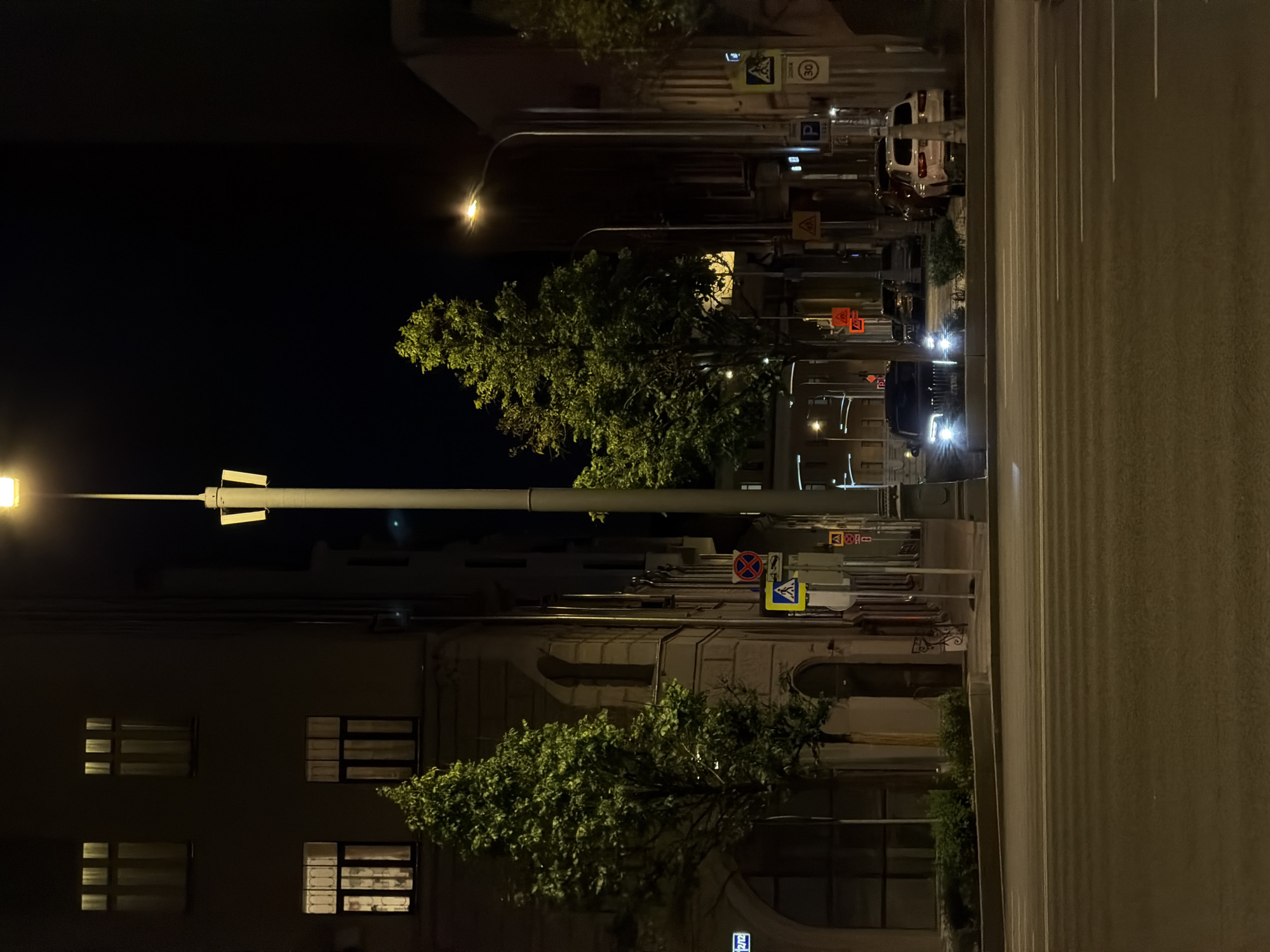 The image captures a nighttime urban scene in Moscow, Russia. A wide street dominates the foreground, illuminated by streetlights. A building with lit windows stands on the left side of the street, partially obscured by trees. Streetlights with security cameras are visible along the sidewalk. Traffic and street signs indicate the presence of moving vehicles and pedestrian crossings. Parked vehicles are seen further up the road. The sky is dark, indicating it is late at night.