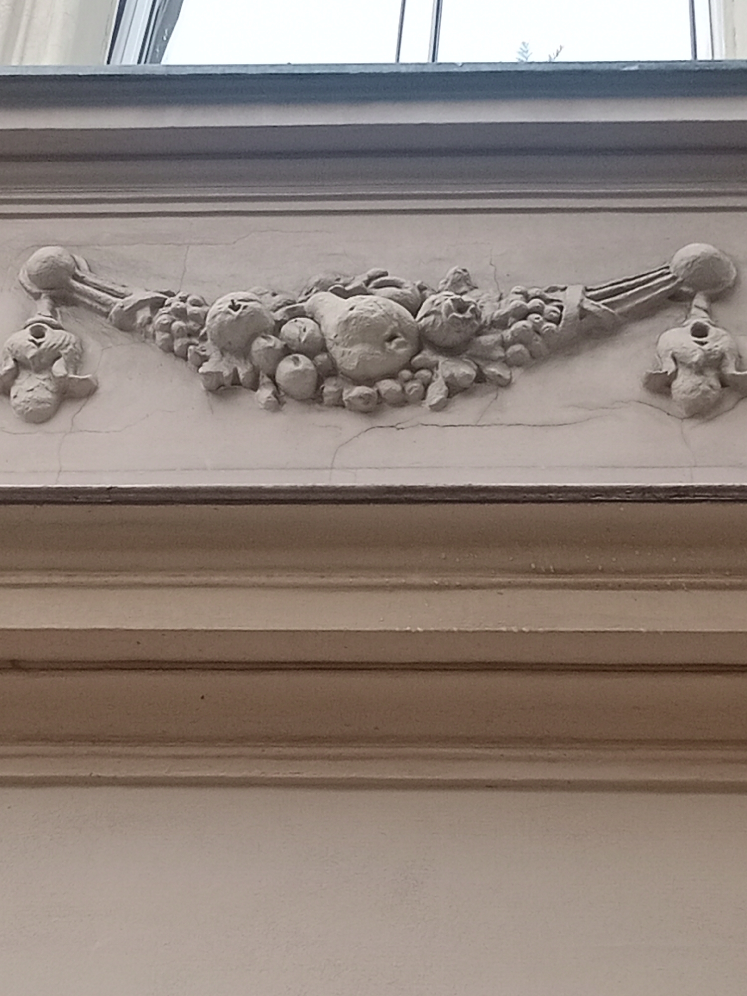The image shows a detailed architectural element, specifically a plaster frieze, above a wooden paneling. The frieze features a central garland of fruit and foliage, flanked by two decorative figures. The fruit appears to include apples and possibly pomegranates, rendered with realistic textures. The foliage is dense and intertwined with the fruit. The decorative figures on either side are stylized, with small, round heads and slender bodies, possibly representing putti or mythological figures. The entire frieze is made of light-colored plaster, showing some signs of age and minor cracking. The frieze is mounted on a textured, pale wall. Below the frieze, there is a prominent horizontal wooden structure with multiple layered moldings, painted in a warm, neutral tone. Further down, the wall is a plain, lighter color. Above the frieze, a window is partially visible, reflecting light and showing a glimpse of greenery outside. This architectural detail is located in the Bydgoszcz City Hall, at Wolności Square 1 in Bydgoszcz, Poland.