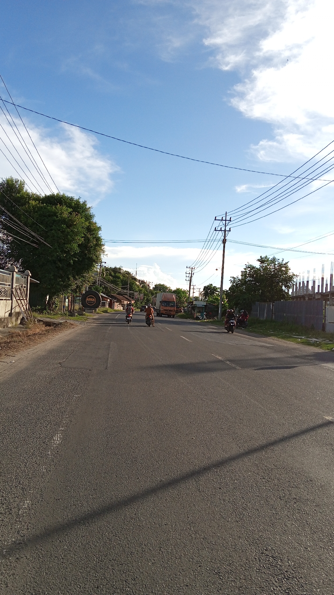 The image depicts a street scene in Cotbadatunong, Indonesia, under a clear blue sky with scattered white clouds. The perspective is from ground level, looking down a paved road. On the left side of the road, a large, leafy green tree stands beside a fence. Further down the road, there are several buildings and a large circular sign with the word "AREA" in it. A few motorcycles are visible on the road, with riders. In the distance, a large orange truck is driving on the road, and on the right side, there are more buildings, including some under construction with scaffolding. Electrical poles and wires are strung across the sky, a common sight in urban and semi-urban environments in Indonesia. The overall impression is of a typical day in a bustling Indonesian town.