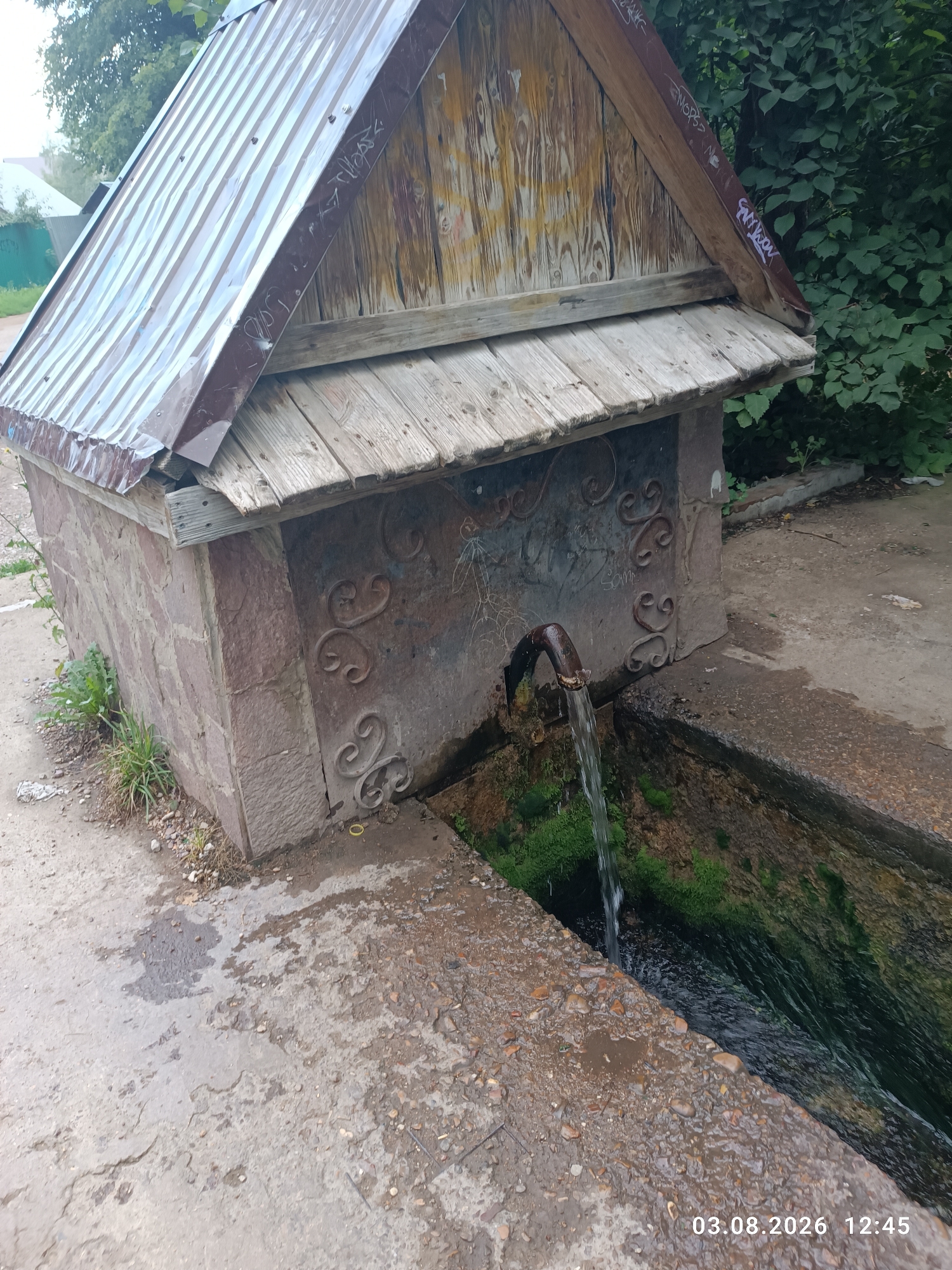 The image depicts an outdoor water source, likely a well or spring, situated in a paved area. The structure consists of a stone base supporting a wooden roof with corrugated metal sheeting. A dark metal pipe protrudes from the stone, from which water is actively flowing into a narrow, overgrown channel. The surroundings include vegetation and some debris on the ground. The timestamp "03.08.2026 12:45" is visible in the lower right corner. The context provided identifies this location as Sterlitamak, Russia. There are no people visible in the image.
