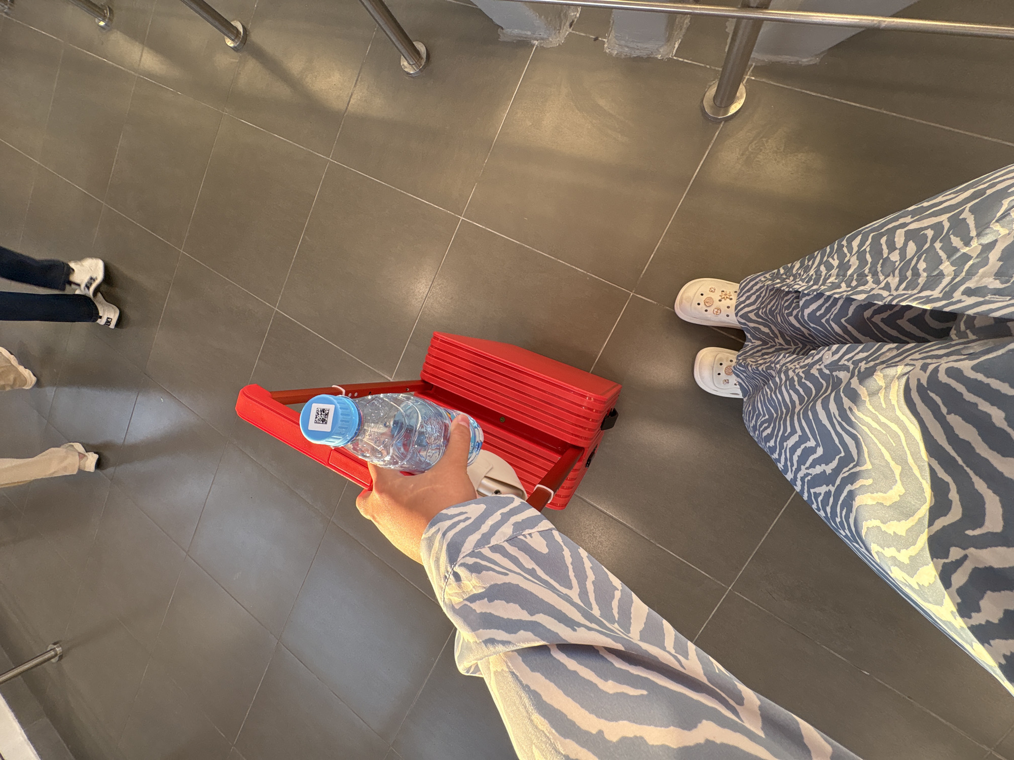 In a high-angle, close-up shot of feet and a hand on a tiled floor, a person wearing a blue and white zebra-print maxi dress and white clogs decorated with shell-like patterns stands on dark gray square tiles. The person's left hand, adorned with a blue and white zebra-print long-sleeved shirt, holds a clear plastic water bottle with a blue cap. The bottle features a QR code on its label. The bottle is positioned above a small, bright red rolling suitcase with a silver handle. The background shows more of the dark gray tiled floor and appears to be an indoor public space, possibly a waiting area or hallway. In the mid-ground, two other individuals are visible from the knees down, one wearing beige pants and white sneakers, and the other wearing dark blue pants and white sneakers. Metal railings and supports are visible on the right side of the frame. The location is identified as being in Matveevka, Russia.