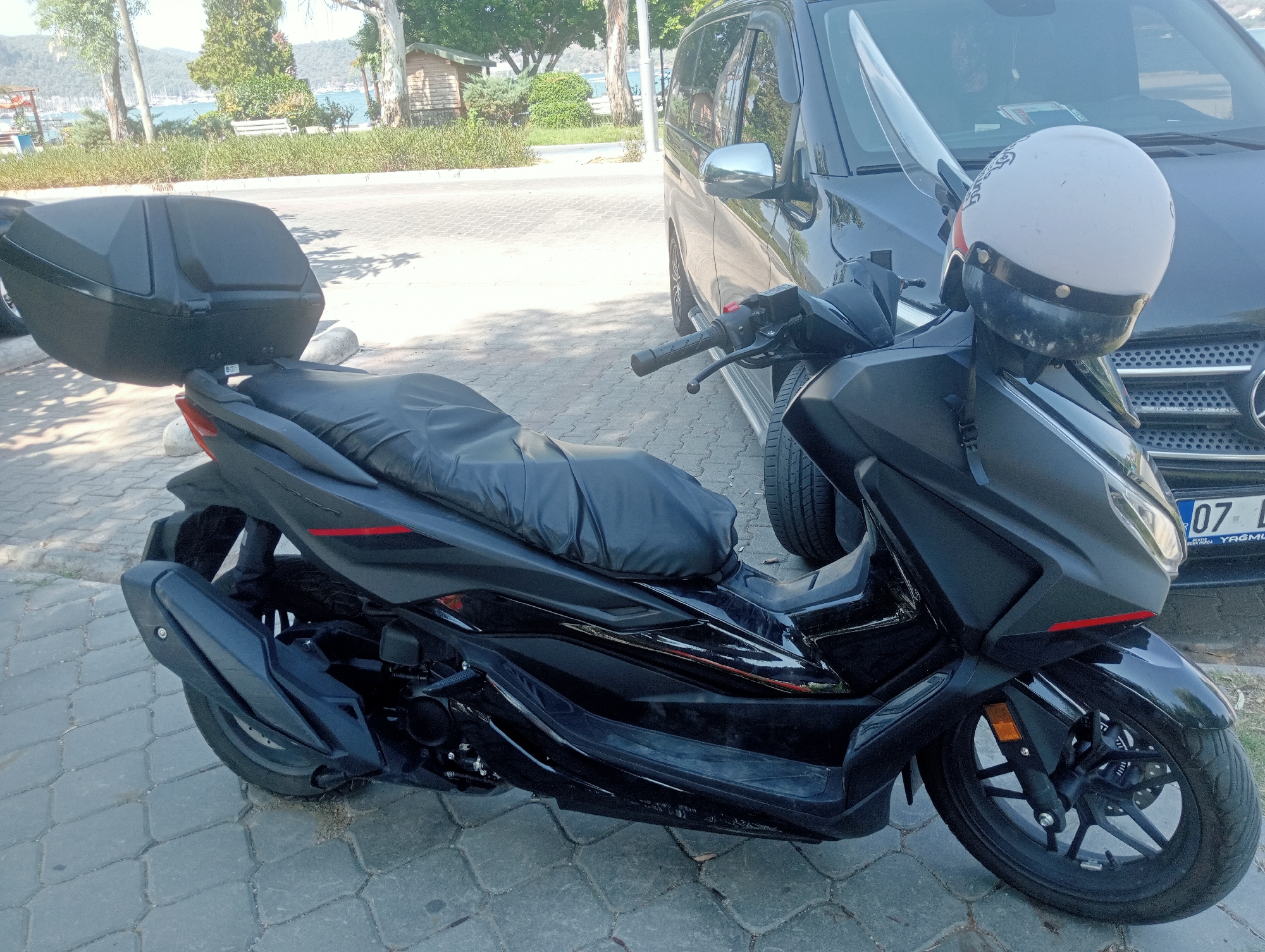 A black scooter with a black seat cover and a black top case is parked on a paved area next to a dark-colored van. A white helmet with a black visor and red accents is resting on the handlebars of the scooter. The van has a license plate that reads "07 L." The background shows trees, a body of water with sailboats, a wooden structure, and a white bench. The overall scene suggests a sunny day in a coastal city. This image was taken in Fethiye, Turkey.