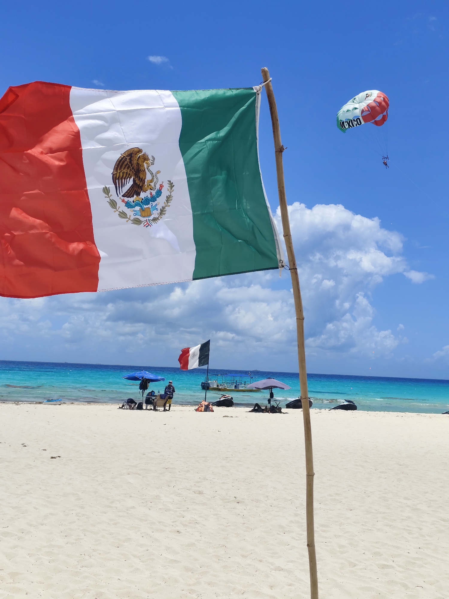 The image shows a beach scene in Playa del Carmen, Mexico. A Mexican flag is prominently displayed in the foreground. In the background, the turquoise water of the Caribbean Sea meets the white sand beach. Several people are relaxing on the beach, some under umbrellas. A parasail with "Mexico" written on it is visible in the sky, pulled by a boat. There is another, smaller Mexican flag further down the beach. Jet skis are parked on the sand near the water's edge.