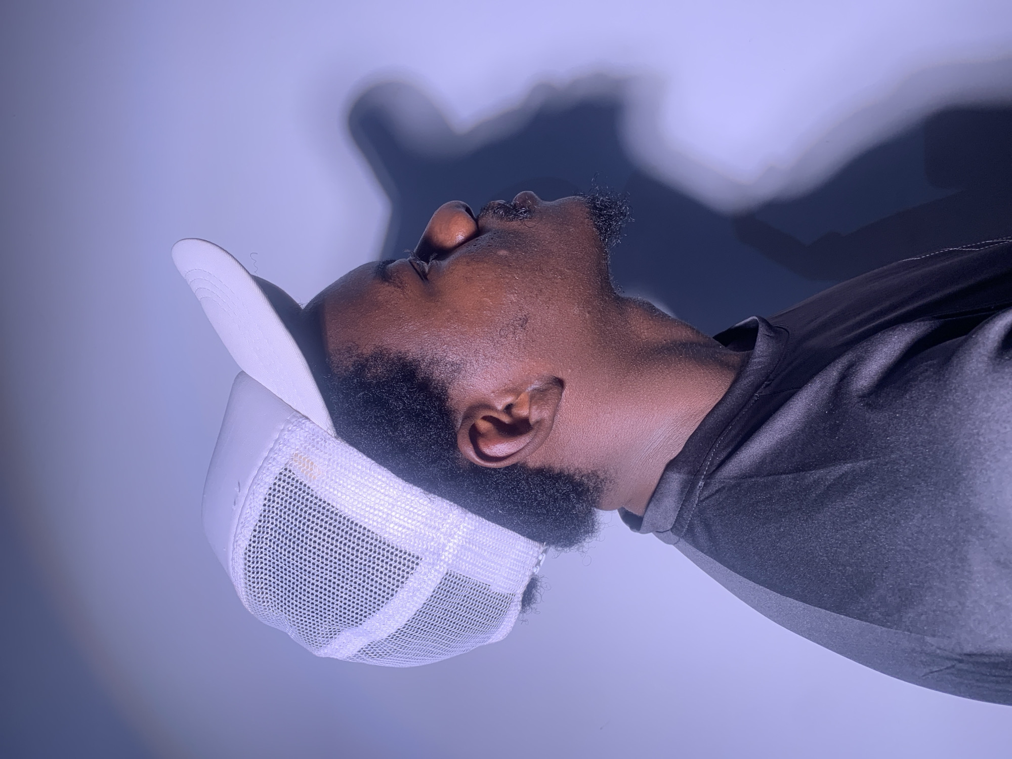 A man wearing a white mesh trucker hat and a dark gray t-shirt is pictured in profile against a muted purple background. He has dark skin, short black hair, and a small goatee. A shadow of his head is cast on the wall behind him. The lighting suggests an indoor setting, possibly a studio or a room with controlled lighting. There are no visible elements that indicate a specific city or location.