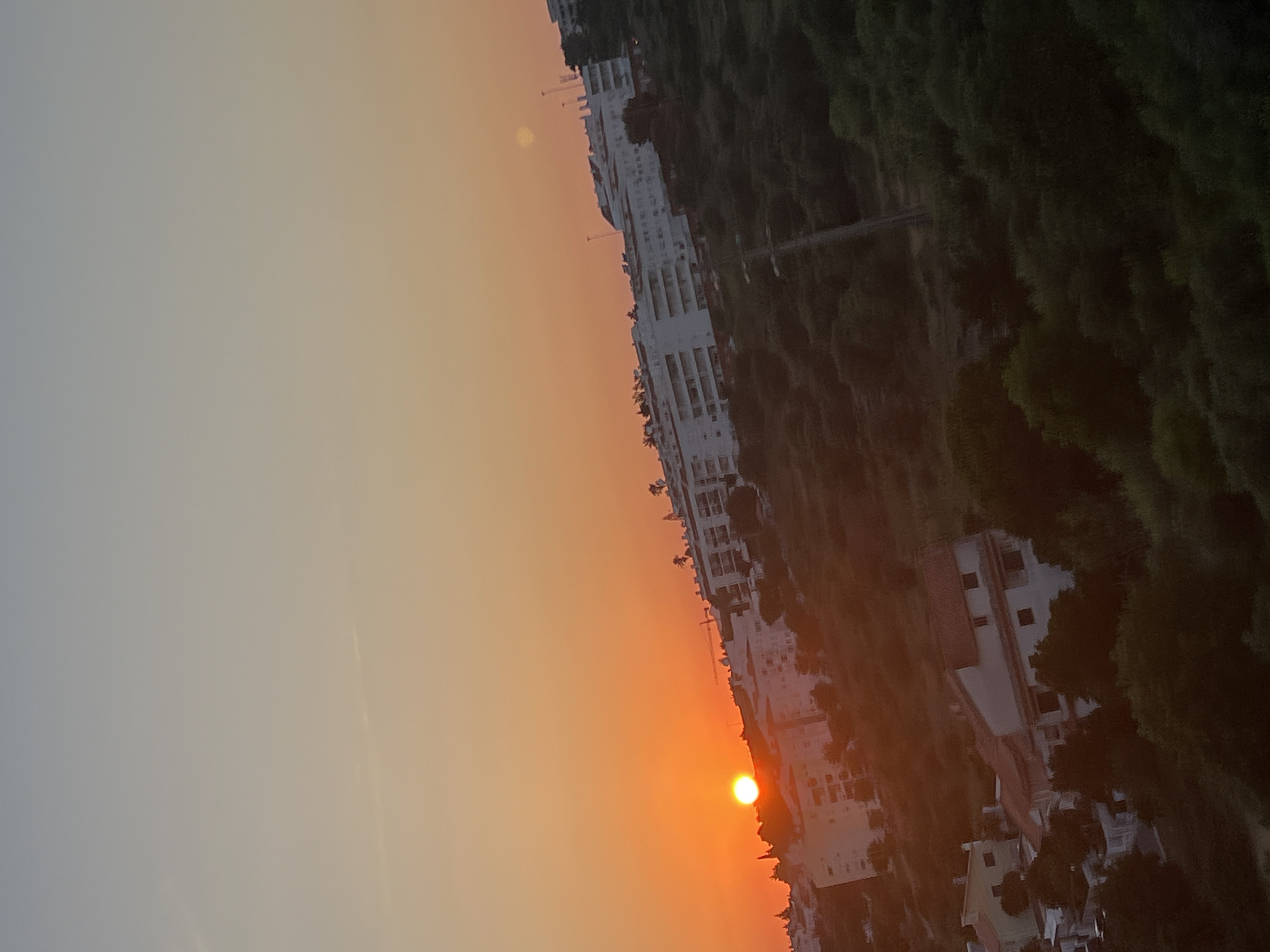 An extreme wide-angle, low-angle shot captures a dusky sunset over a hilly urban landscape in Cascais, Portugal. The sky transitions from a soft blue at the top to a vibrant orange near the horizon, where a bright, orange sun is visible. Dominating the mid-ground are several white apartment buildings with visible balconies and windows, some appearing to be multi-story structures. These buildings are nestled amongst lush, dark green trees and foliage that cover the undulating terrain. In the foreground, smaller residential buildings with red-tiled roofs are scattered amongst the trees. A faint construction crane is visible in the distance, behind the apartment buildings. The scene is a mix of natural landscape and human habitation, bathed in the warm, fading light of dusk.