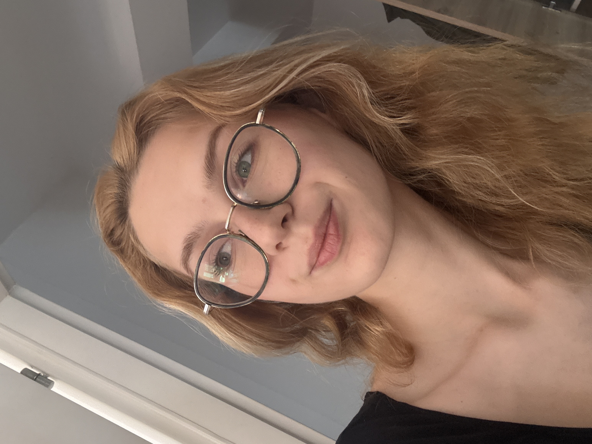 A young woman with blonde hair and glasses is shown from the chest up in a selfie. She is looking to her left, and her mouth is slightly open in a smile. She is wearing a black top. The background is out of focus and appears to be an indoor setting with a light-colored wall and a doorway. There is no indication of the city of Kragujevac, Serbia.