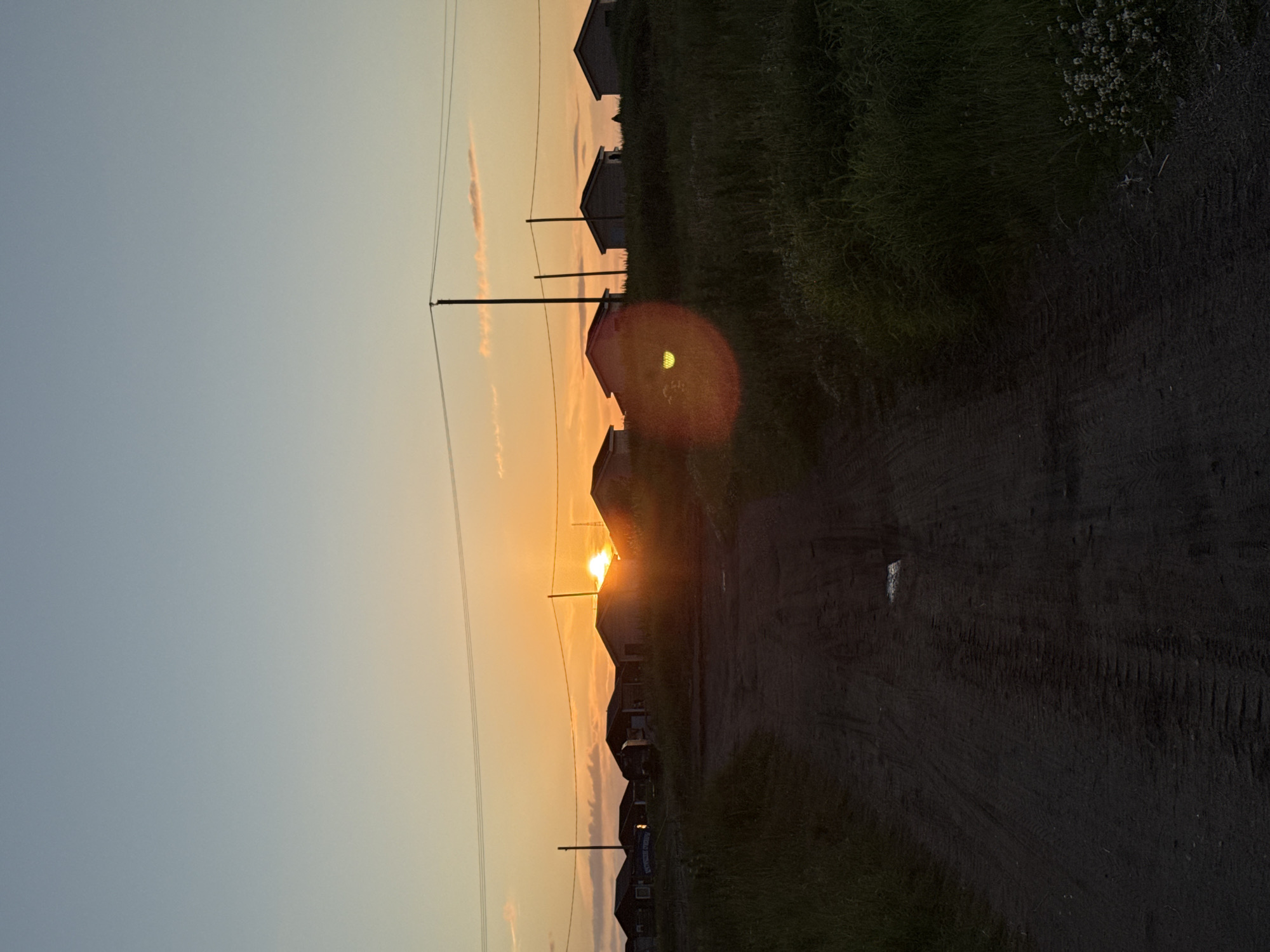 The image depicts a silhouetted landscape with a dirt road leading towards a line of houses in the background. The sun is setting, casting an orange glow across the sky and illuminating the horizon. Power lines and utility poles are visible against the sky. The foreground shows a dark, muddy dirt road with tire tracks, bordered by tall grass and some small white flowers on the right. The houses appear to be simple, residential structures, some with dark roofs. The overall scene suggests a rural or semi-rural setting in Teriberka, Russia, during twilight. There are no people or animals visible in the image.