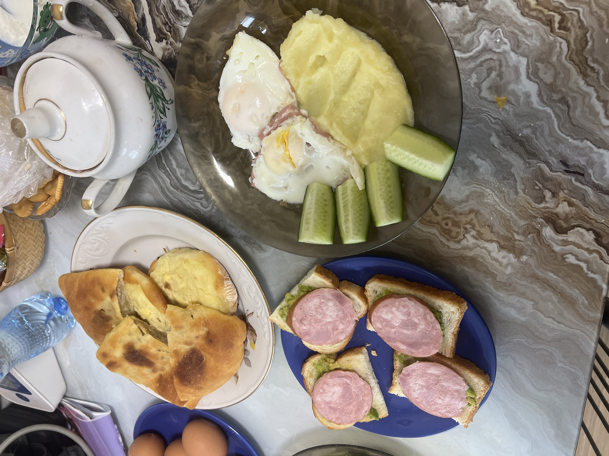 This image presents a varied meal spread across a marbled tabletop, suggesting a casual breakfast or brunch setting in a home, potentially in Perm, Russia. No people are visible.
A dark, smoky glass plate in the upper right quadrant holds a serving of mashed potatoes, two fried eggs with visible yolks, and three thick slices of fresh cucumber.
To the left, a white plate with a decorative gold rim displays a large, golden-brown baked bread or pastry, cut into several pieces, revealing a light-colored filling, possibly cheese or a cream-based mixture. Nearby, a clear plastic bottle of water and a white teapot with blue floral patterns and gold accents are visible. A partially obscured bowl next to the teapot contains what appears to be a dairy product like sour cream or yogurt. A plastic bag with crackers is also present.
In the lower right, a bright blue plate holds four toasts, each covered with a green spread, likely avocado or guacamole, and topped with a large, round slice of pink cooked sausage or bologna.
The bottom left corner reveals a blue bowl containing several raw brown eggs. The scene conveys a hearty and diverse meal, typically served in the morning or early afternoon. There is no discernible visible text.