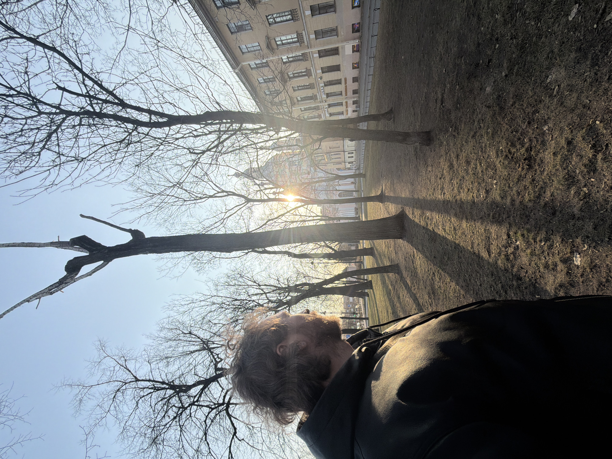 The image presents a low-angle view of a man with wavy brown hair and a beard lying on the ground, possibly in a park or green space. He is wearing a black jacket. Above him, there are several bare trees with intricate branch patterns against a pale blue sky. The sun is shining through the trees, creating streaks of light.
In the background, there is a building with multiple windows, indicating an urban setting. Based on the architecture and the presence of parks, it is very likely that this image was taken in Saint Petersburg, Russia. The overall scene conveys a sense of tranquility and a moment of contemplation.