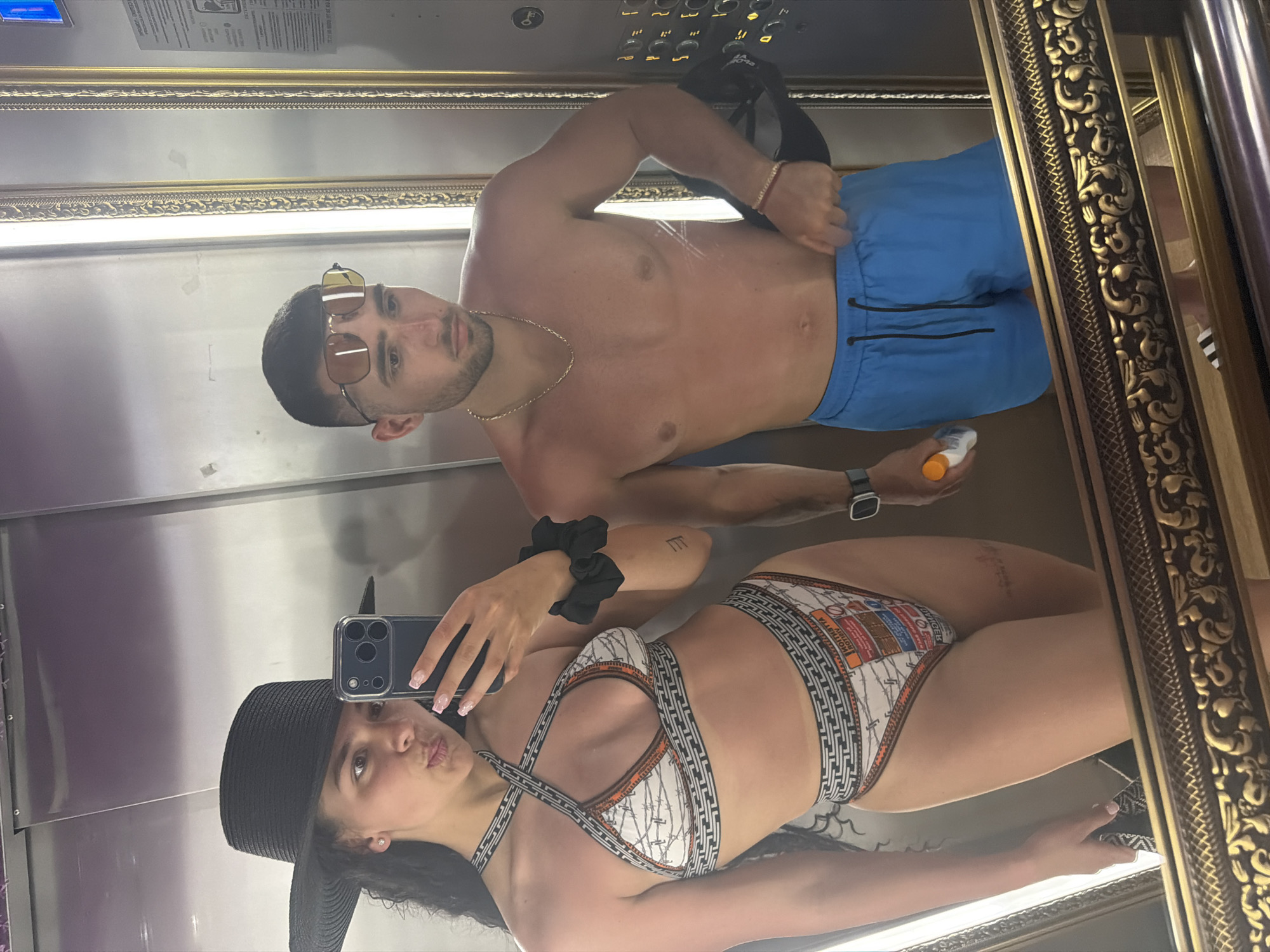 In an elevator with ornate gold trim, a young couple poses for a mirror selfie. The man, shirtless and wearing blue swim trunks, stands behind the woman. He has short dark hair and is wearing aviator sunglasses and a gold chain necklace. A thin gold bracelet adorns his wrist. He holds a bottle of sunscreen in his right hand, and a black baseball cap is visible on the back of his head. The woman, in the foreground, is wearing a black sun hat and a patterned bikini. Her bikini top features a white background with black, orange, and geometric patterns, and criss-cross straps. The bikini bottoms have a similar design. She has dark curly hair and is holding her phone to take the picture. Her nails are painted a pale pink. A black scrunchie is visible on her left wrist. The mirror's reflection also shows elevator buttons and a partial view of what appears to be a beach or resort setting. The overall mood appears casual and relaxed, possibly during a vacation. The image seems to be taken during the day, given the lighting.