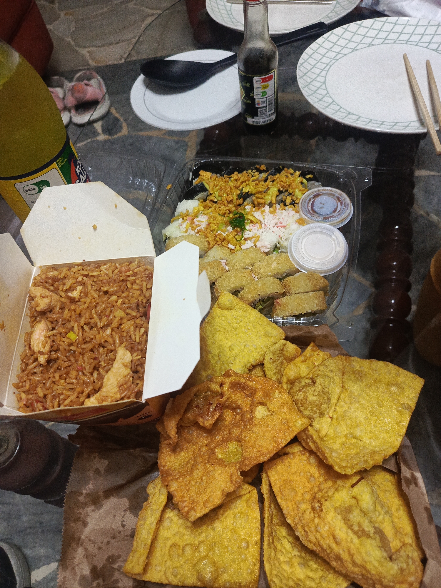 An overhead shot captures a dimly lit table displaying a meal. In the foreground, a pile of golden-brown fried wontons and a carton of chicken fried rice sit on a brown paper liner. To the right, a clear plastic container holds sushi rolls topped with shredded vegetables and white sauce. A small bottle of soy sauce and two small white dipping sauce containers are positioned above the sushi. In the background, a yellow bottle of drink and a black spoon rest on a white plate. The scene appears to be set indoors on a tiled surface, possibly a dining table in a home in Guayaquil, Ecuador.