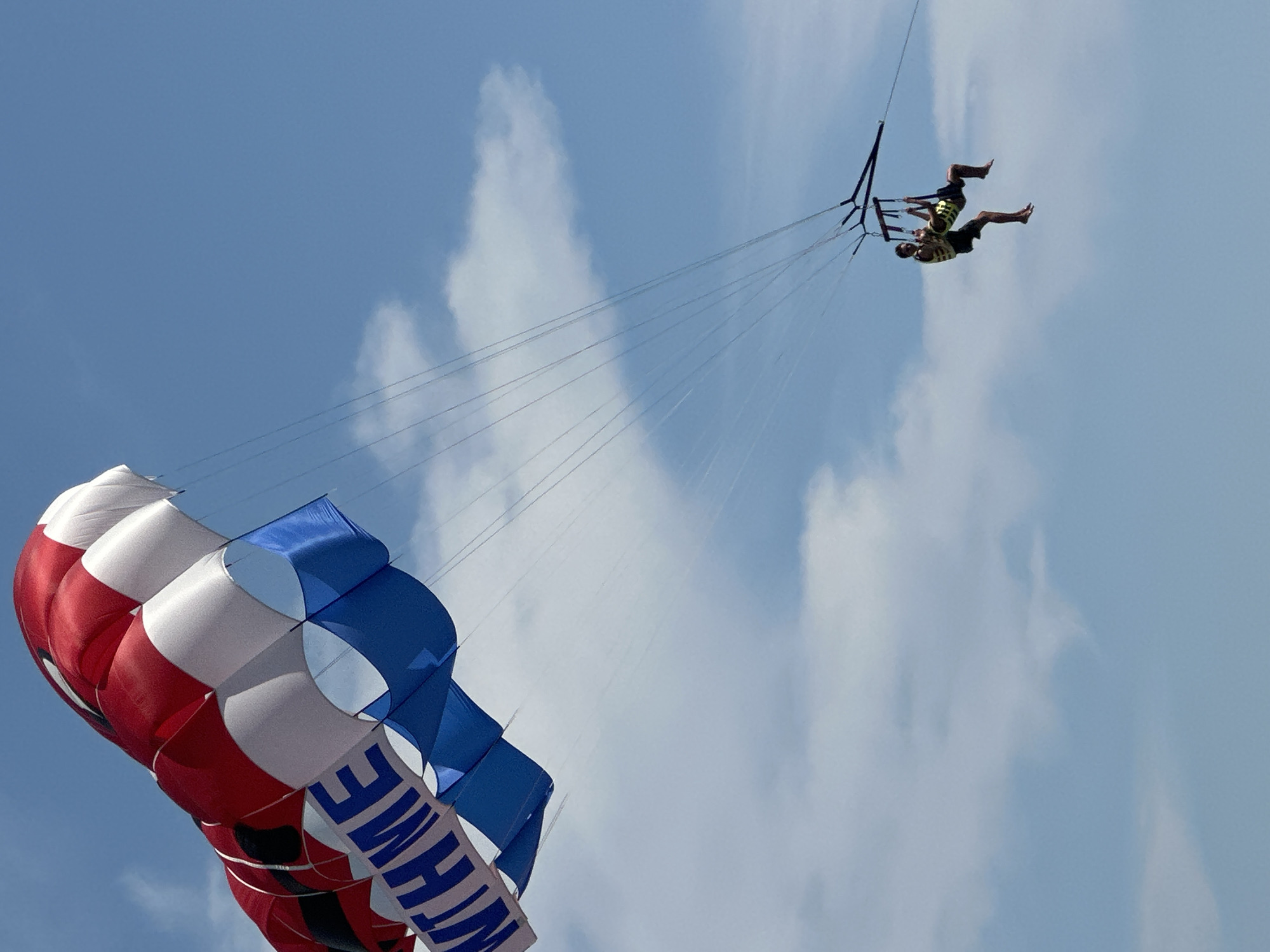 An individual is parasailing in the sky above the city of Budva, Montenegro. The parasail is red, white, and blue and has the word "WITH ME" written on it in blue letters. The person is wearing a yellow life vest and dark shorts. The sky is clear and blue with a few white clouds. The image was taken from below the parasail, looking up.