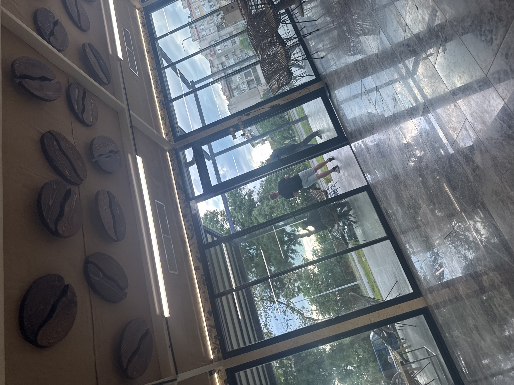 The image shows the interior of a cafe. On the left wall, there are decorative wooden coffee beans arranged vertically. The right side of the image displays a marble-tiled floor reflecting the scene outside. The cafe has large glass windows providing a view of the outside. Through the windows, a cityscape with buildings is visible, as well as trees. Two people are seen walking along the sidewalk outside the cafe. Chairs and tables are positioned near the windows. Based on context, this is likely a cafe in the city of Yambol, Bulgaria.