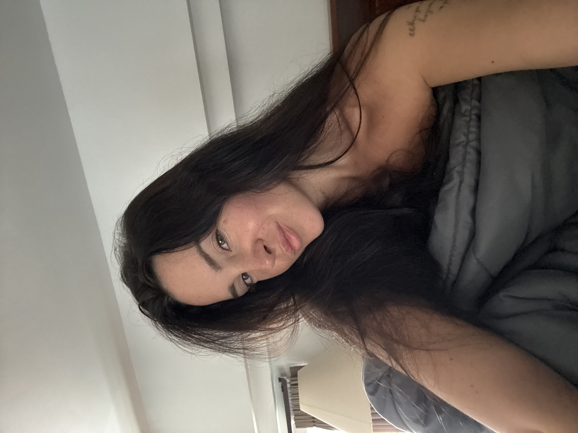 The image is a close-up, vertically oriented selfie of a young woman with long, dark hair. She is lying down, partially covered by a gray blanket, and appears to be in a relaxed or intimate setting, possibly a bedroom. Her head is tilted, and she is looking directly at the camera with a soft, contemplative expression. Her skin is fair, and she has noticeable freckles on her nose and cheeks. A tattoo is visible on her upper arm. The background is blurred, with hints of a wall and possibly a bed frame. The lighting suggests it could be morning or evening, given the soft, diffused light. The overall mood is personal and intimate. There is no discernible text in the image. The provided location context of North Kuta, Indonesia, does not offer specific visual cues within the image itself to confirm or deny this.