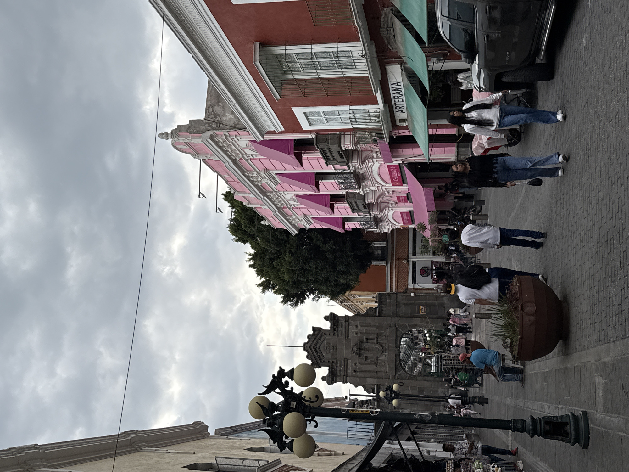The image depicts a bustling street scene in a historic urban setting, consistent with Puebla, Mexico, under an overcast sky. The perspective is from a pedestrian level, looking down a paved street lined with buildings and people.
On the left side of the street, a striking multi-story building features a vibrant pink facade with intricate white architectural detailing, decorative balconies with dark railings, and several pink awnings. Above it, another building with a reddish-brown exterior and white-framed windows secured with iron bars is visible. A large, dark green tree stands further down the street on this side.
The street is paved with dark, possibly cobblestone or interlocking brick, material. On the right side, a dark-colored pickup truck is parked. Numerous pedestrians are present; some are walking, while others are standing or observing. They are casually dressed in items like jeans and shirts. A large, dark brown planter with green foliage is situated on the right side of the walkway.
In the foreground on the left, an ornate black streetlight with multiple spherical, pale yellow globes is prominent. Another similar streetlight is visible further down the street. In the distance, a grand stone building with an elaborate archway and facade adds to the historical ambiance. The sky is predominantly cloudy, suggesting a day without direct sunlight.
Visible text includes "ARTERAMA" on a white sign mounted under a green awning. A partially obscured red sign further down reads "CARNICERIA S" (likely part of "Carnicería S.A."). The overall scene presents a lively urban environment with a blend of historical architecture and contemporary activity.
