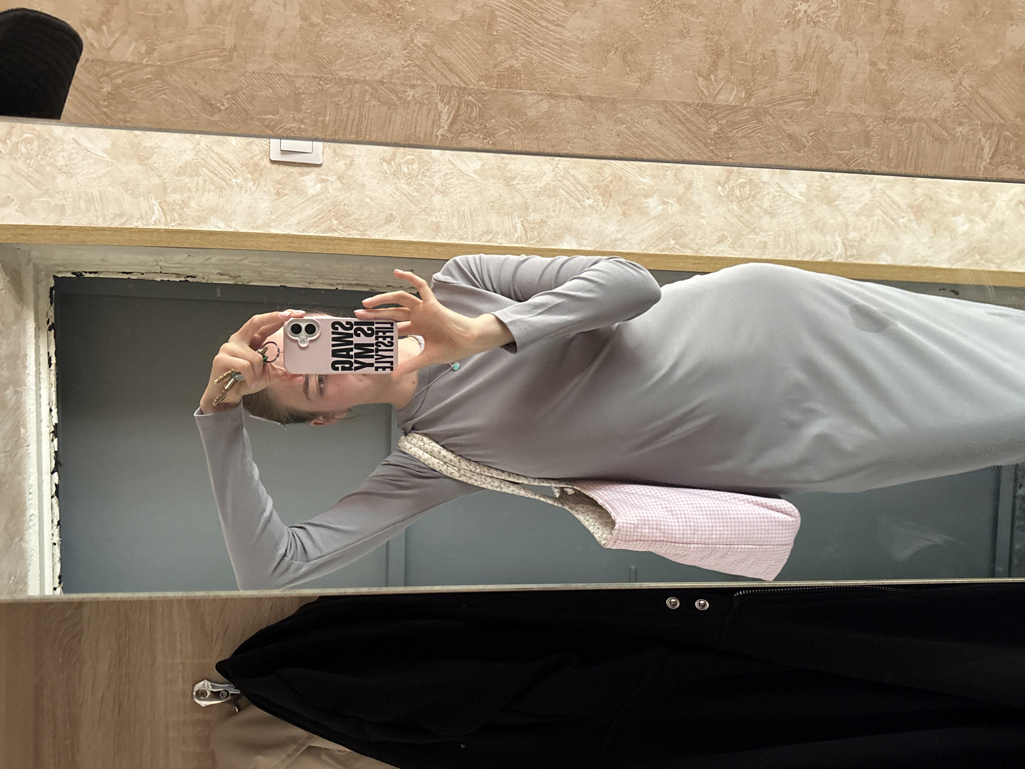 A person is taking a mirror selfie in Perm, Russia. The individual is wearing a grey long-sleeve dress and has a pink and white checkered handbag. A black jacket is visible hanging below the mirror. The mirror reflects a wall with textured beige wallpaper and a light switch. The mirror itself has a silver frame. The person is holding a pink phone with a case displaying skull designs and text "SWAG MY LIFE". The person is wearing multiple rings.