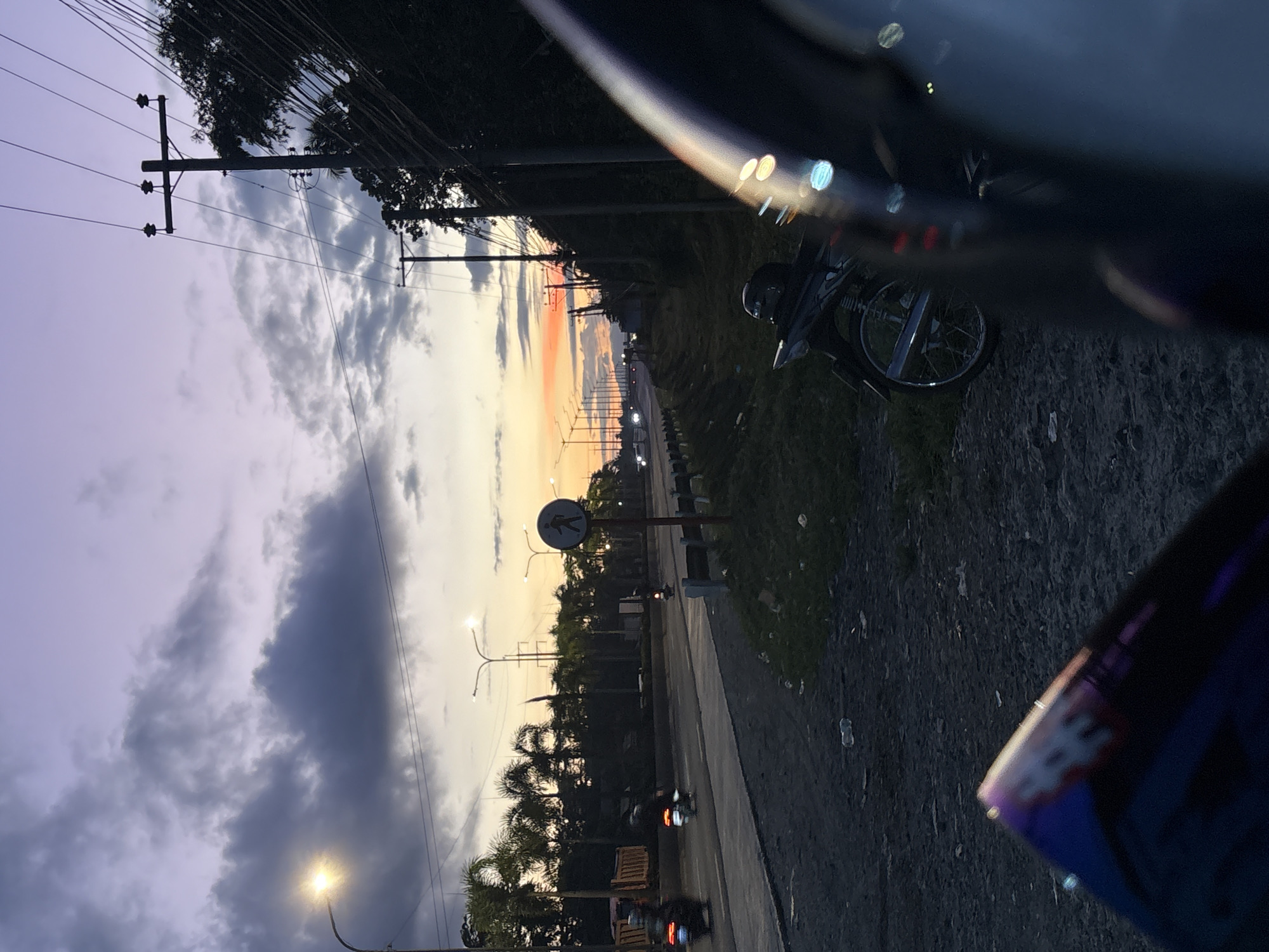 The image depicts a dusk scene on a road in Carmen, Philippines. In the foreground, the edge of a vehicle or motorcycle accessory is visible, partially obscuring the view. To the right, a parked motorcycle with a helmet on its seat is positioned on the gravelly shoulder of the road. The road itself is paved and shows signs of traffic, with streetlights lining its length and a pedestrian crossing sign with a walking figure symbol. Several motorcycles and a truck are visible on the road, some in motion, indicated by blurred taillights. Palm trees and dense foliage line the side of the road, and power lines with poles stretch across the upper portion of the frame. The sky is a gradient of purple and orange, transitioning from twilight hues to a darker evening sky, with scattered clouds. The overall atmosphere is that of a transitional period between day and night in a tropical urban or semi-urban environment.