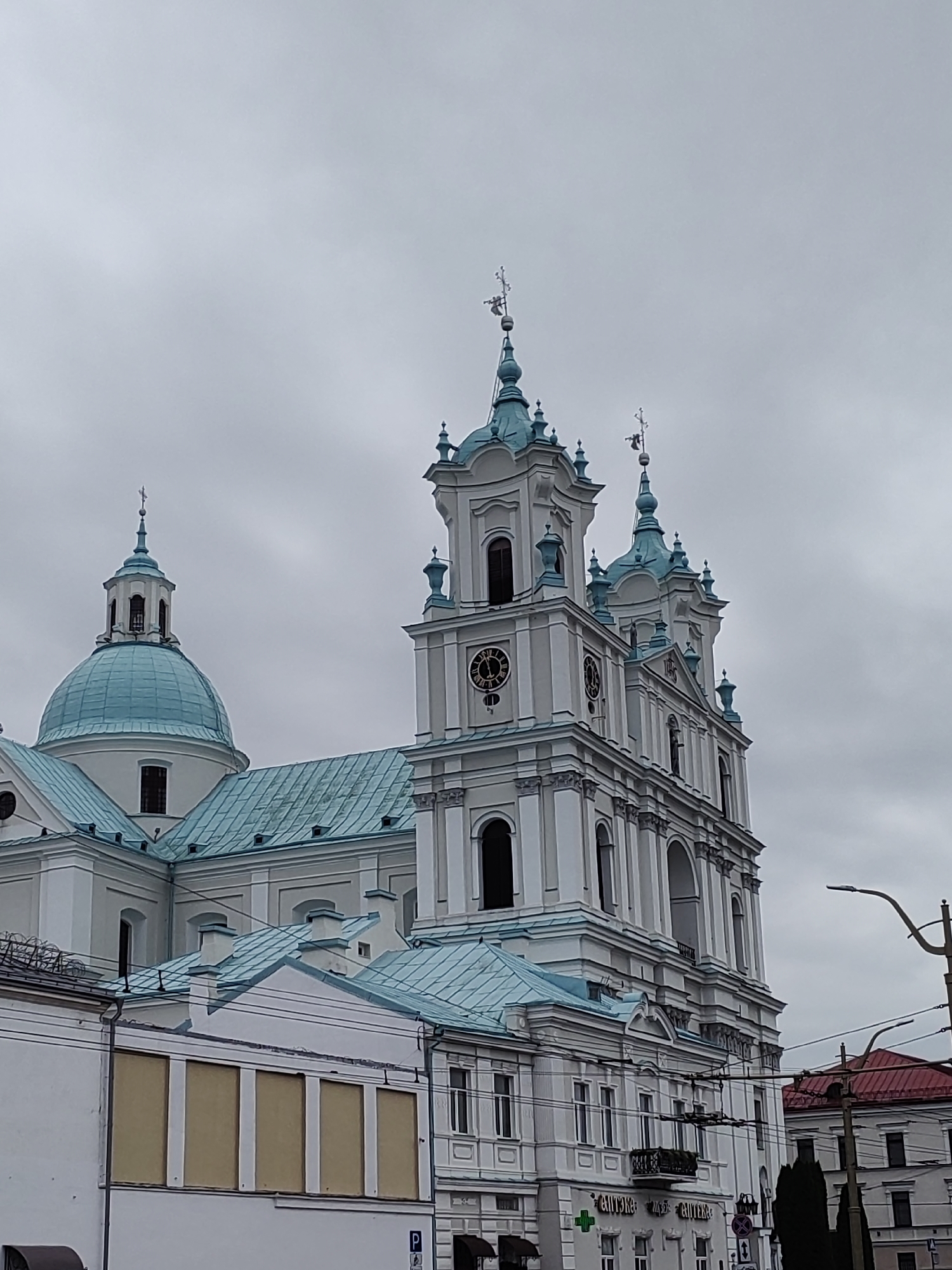 The image captures a view of the Old Catholic Cathedral of St. Francis Xavier in Hrodna, Belarus, on a cloudy day. The grand white facade of the cathedral is adorned with blue-green tiled roofs and spires. The prominent dome on the left is a striking feature, as are the two tall bell towers on the right, one of which displays a clock. In the foreground, a white building with a light brown facade runs along the lower part of the image, with some windows boarded up. Overhead power lines and a streetlamp are visible, indicating an urban setting. A red-tiled roof of another building is visible in the distance to the right. The sky is overcast with a muted, grey hue.