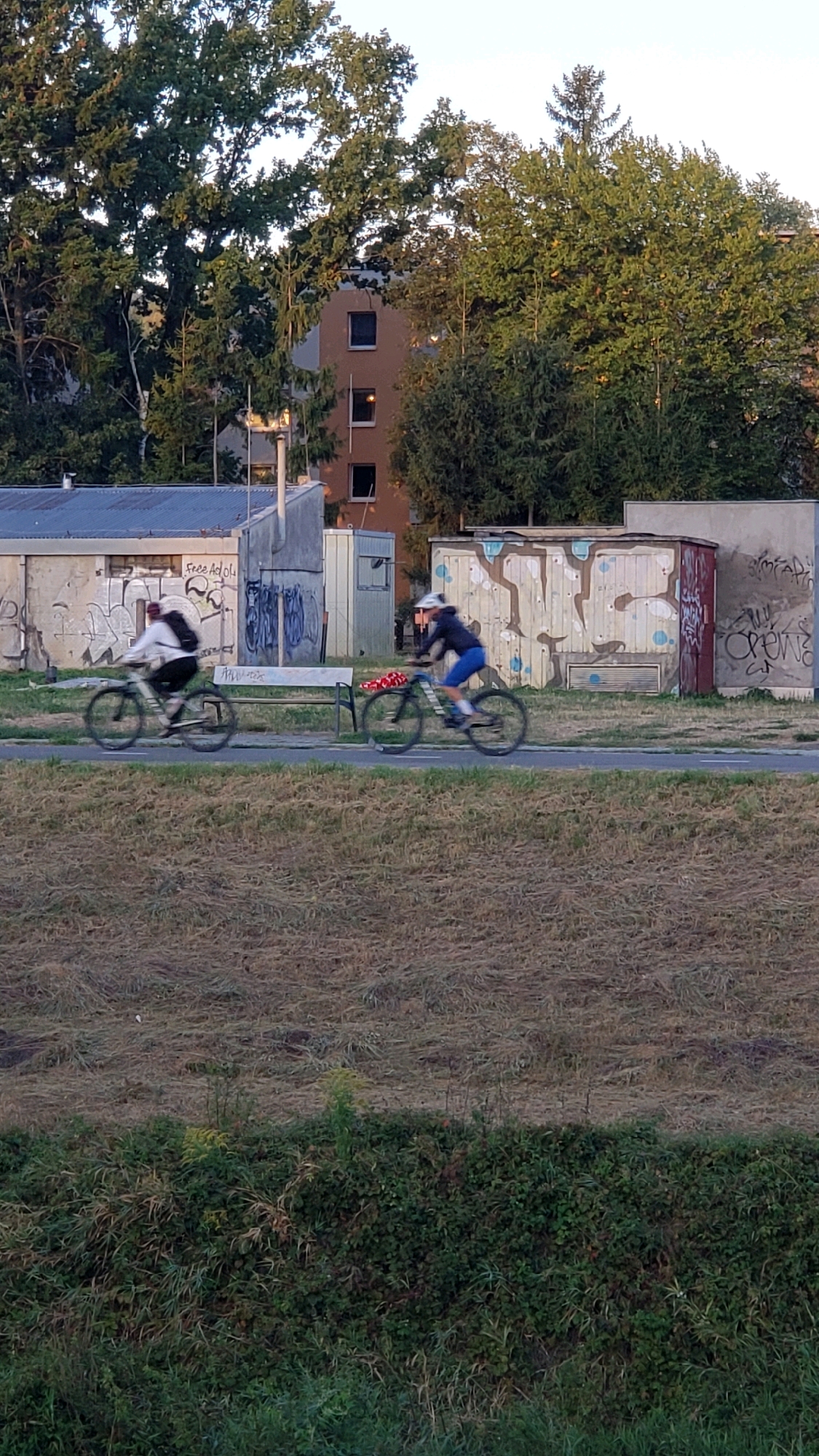 The image shows a blurred view of two people cycling on a path in Prešov, Slovakia. The cyclists are moving from left to right. The person on the left is wearing a light-colored jacket and a backpack, while the person on the right is wearing a dark jacket and blue shorts, with a helmet. Behind them is a row of graffiti-covered sheds or containers, and in the background, trees and a building are visible. The foreground consists of dry grass and a dense line of green vegetation. The sky is clear and light.