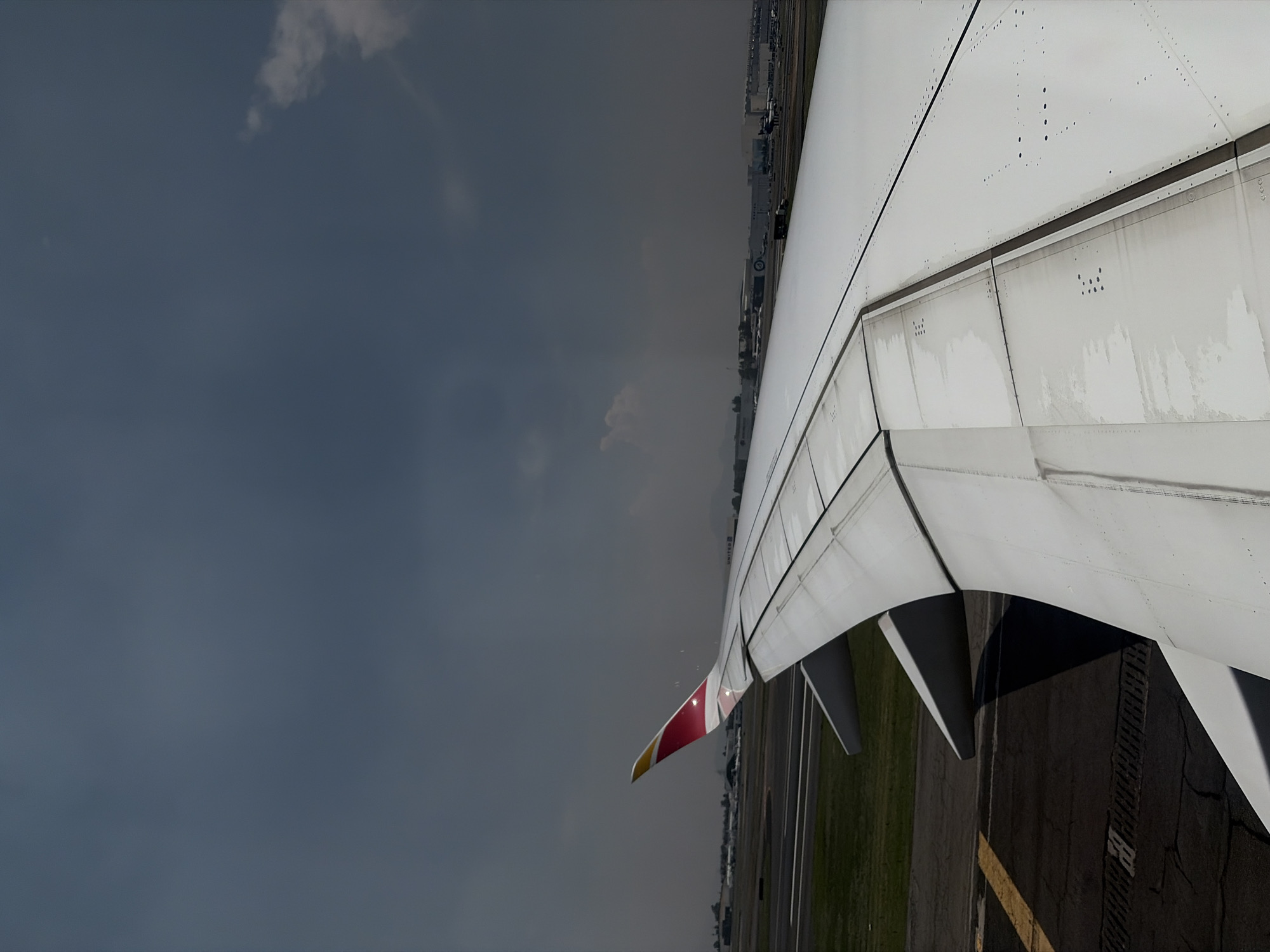 The image shows an airplane wing viewed from inside the aircraft. The wing is primarily white, with a red and yellow tip. The flaps are deployed, suggesting the plane is either taking off or landing.
Outside the wing, the view alternates between a dark, overcast sky and ground features. The ground shows what appear to be airport runways and grassy areas. Buildings can also be seen in the background.