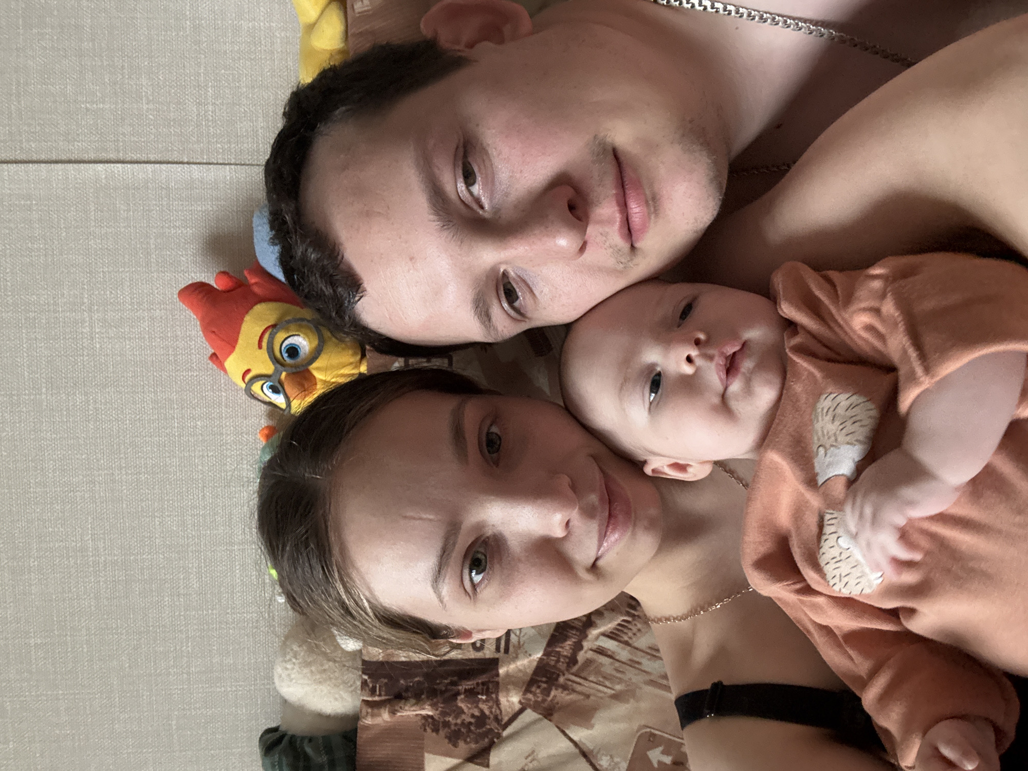 The image is a selfie taken by a family in a horizontal orientation. The family consists of a man, a woman, and a baby. All three are lying down, close together. The man is at the top of the frame, with the woman below him and the baby in between them. All three are looking at the camera. The baby is wearing an orange long-sleeved shirt with a hedgehog design. The woman is wearing a black bra. The man is wearing a silver chain necklace. A yellow stuffed toy with blue goggles is visible to the left of the woman's head. The background includes a beige textured surface, likely a bedspread or blanket. A patterned object is visible in the lower portion of the frame. The location is identified as Chaikovsky, Russia.