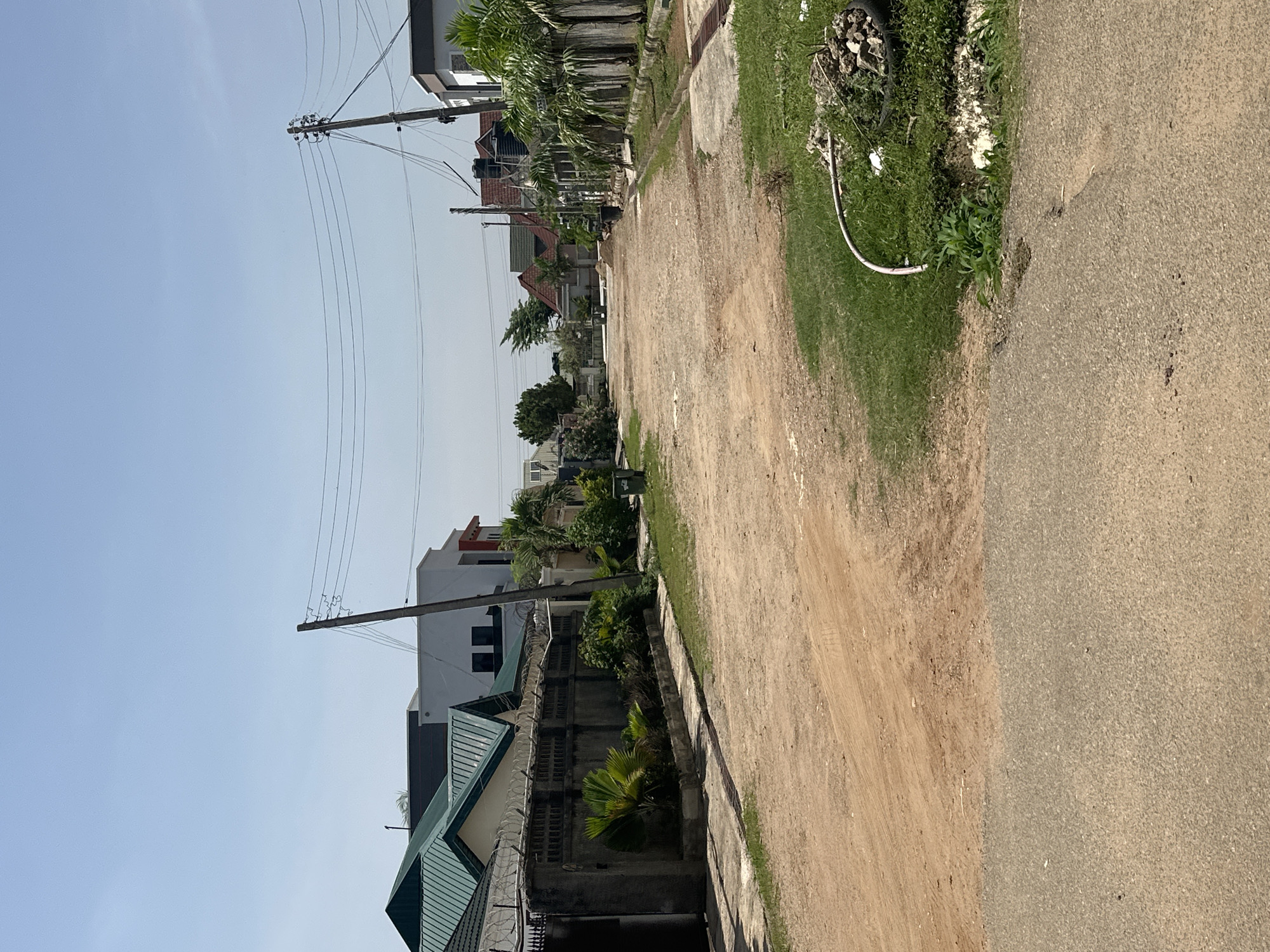 The image depicts a residential street scene in Abuja, Nigeria. The street is unpaved and made of reddish-brown earth, with a paved road visible in the foreground. On either side of the street are houses with various architectural styles, some featuring pitched green roofs and others with flat roofs. Many of the properties are enclosed by high concrete walls topped with barbed wire, indicating a focus on security. Greenery, including trees and shrubs, is present in front of the houses and along the street. Utility poles with overhead power lines are visible along the street. The sky is clear and blue, suggesting a sunny day. There are no people or vehicles visible in the image. The overall impression is of a developing residential area in Abuja.