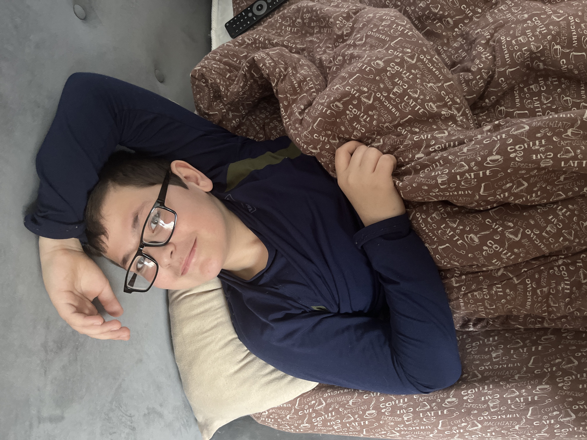 A young boy, likely a pre-teen or teenager, is depicted lying comfortably on a grey upholstered couch or bed. He is positioned on his back, with his head slightly propped on a light-colored pillow, and his left arm resting above his head on the grey surface. He wears black-framed glasses and a dark blue long-sleeved shirt, with a hint of a green or khaki undershirt visible at the collar. His expression is calm and relaxed, featuring a soft, engaging smile directed towards the camera.
He is partially covered by a brown blanket adorned with a repetitive pattern of coffee-related words and illustrations of coffee cups. Visible text on the blanket includes "LATTE," "COFFEE," "MACCHIATO," "AMERICANO," "CAPPUCCINO," "ESPRESSO," and "CAFE." A black remote control is also resting on the blanket in the upper part of the frame. The overall setting is indoors, suggesting a cozy and casual home environment in Vesyoloye, Russia. The lighting appears soft, typical of an interior shot, and the scene conveys a sense of comfort and repose.